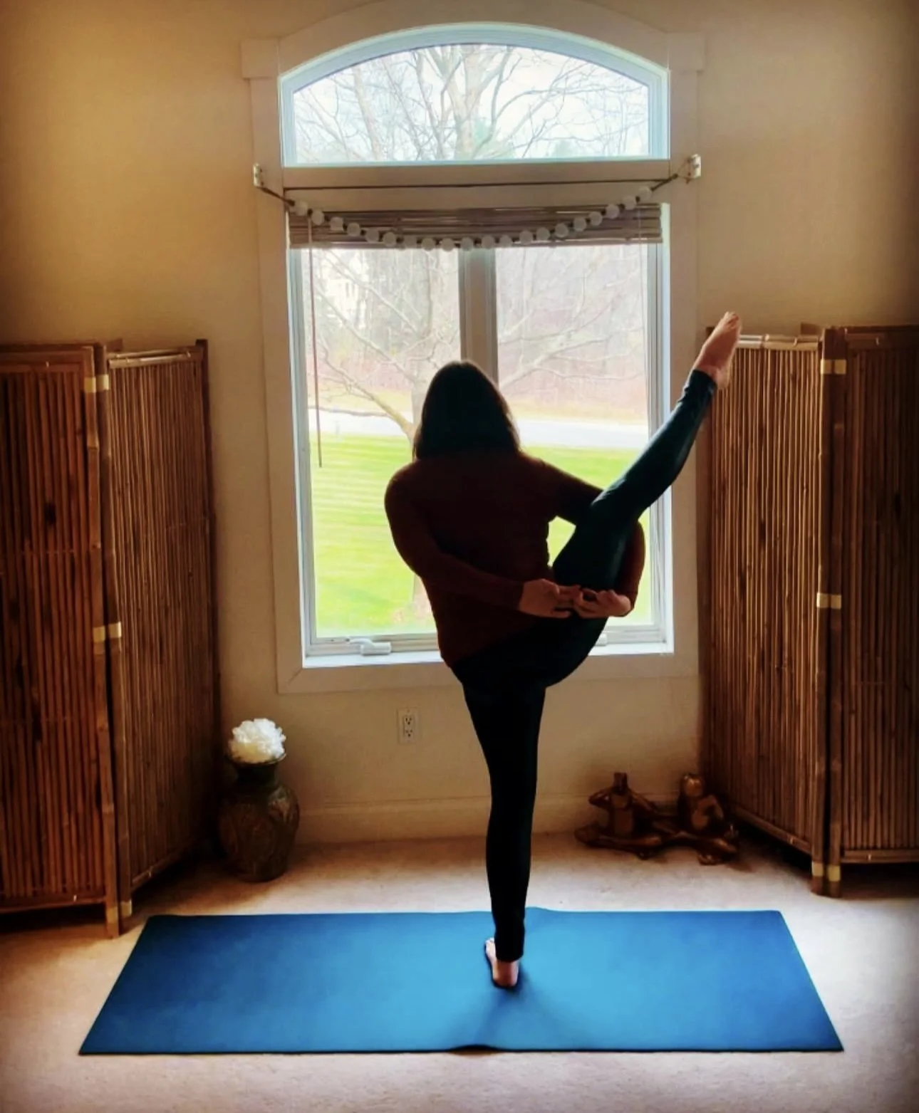 A woman practicing yoga in a room with bamboo room dividers, a blue mat, and a large window showing trees outside. She is standing on one leg while holding the other leg extended high.