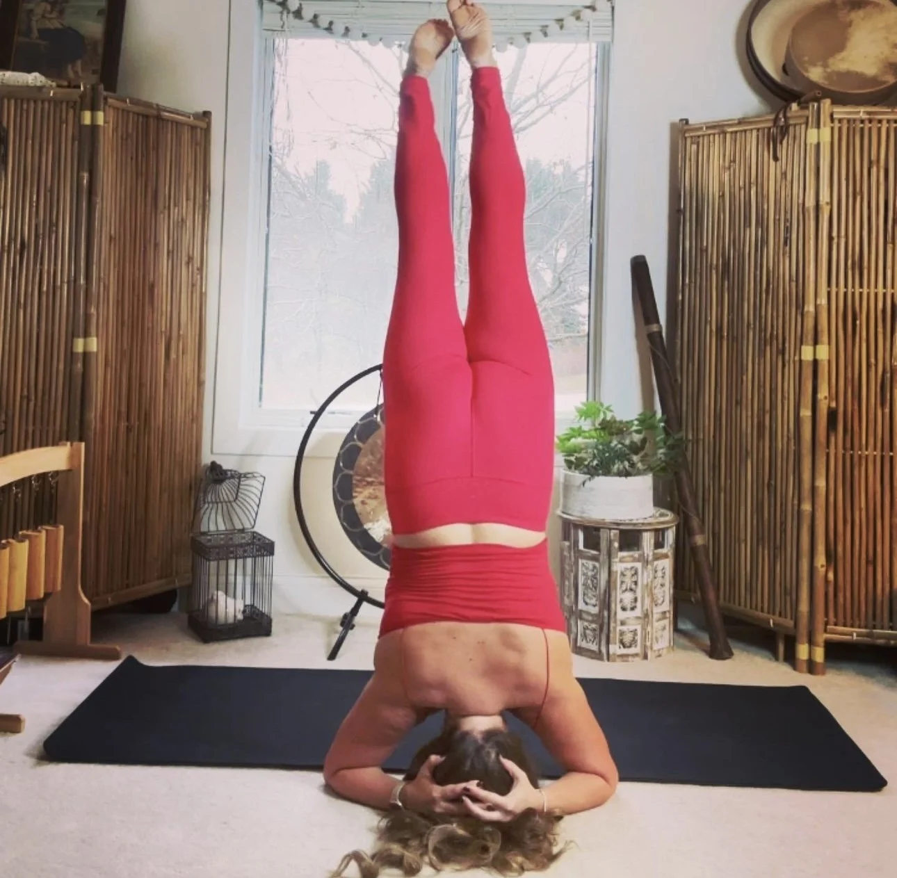 A woman practicing yoga indoors, balancing in a headstand pose on a black yoga mat. She is wearing red leggings and a matching red top, with her hands clasped around her head. The room has wooden folding screens, a small table with plants, a drum, an