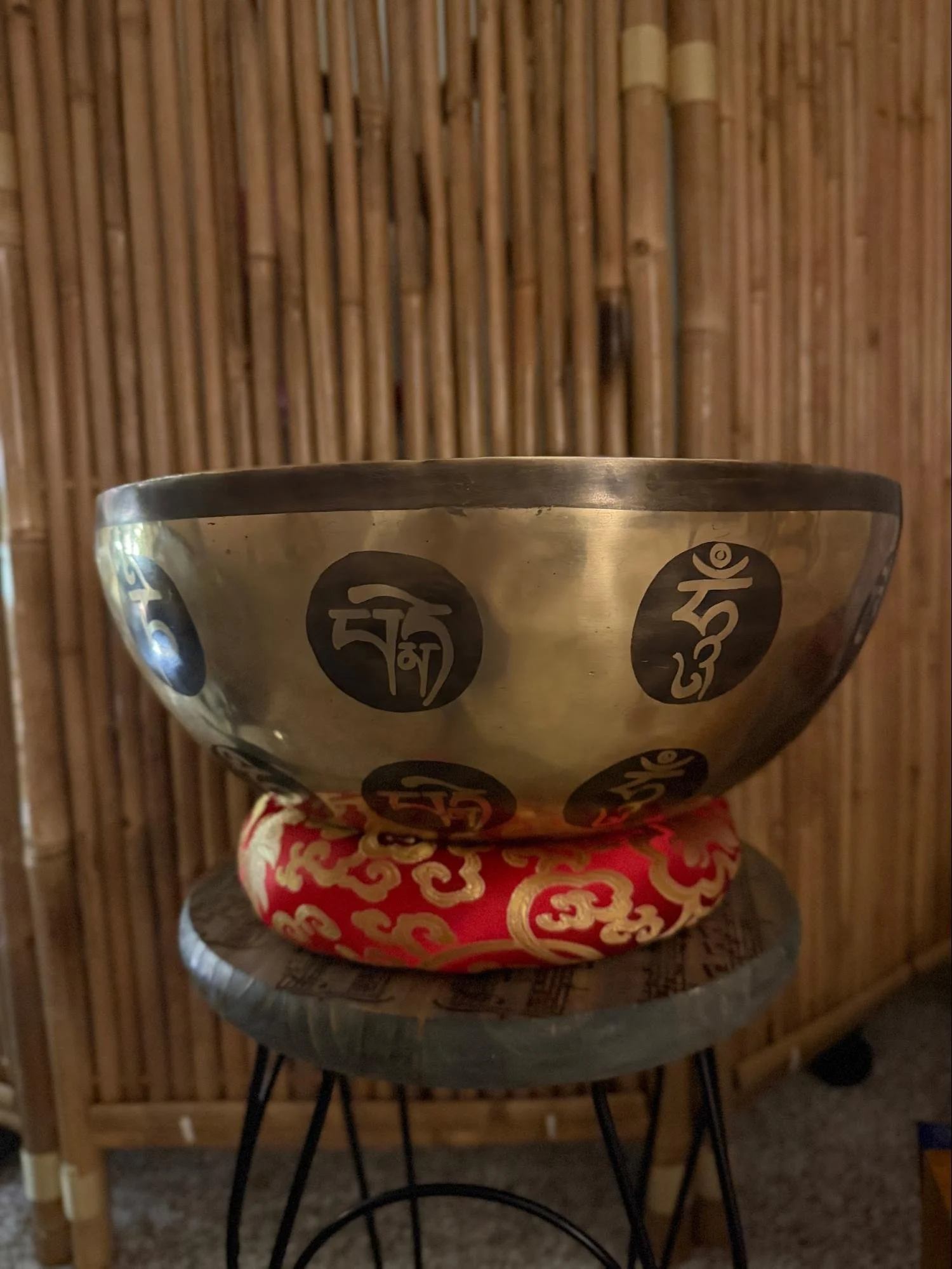 A brass singing bowl with black symbols on its side, resting on a red and gold patterned cloth on a small round table, against a bamboo wall background.