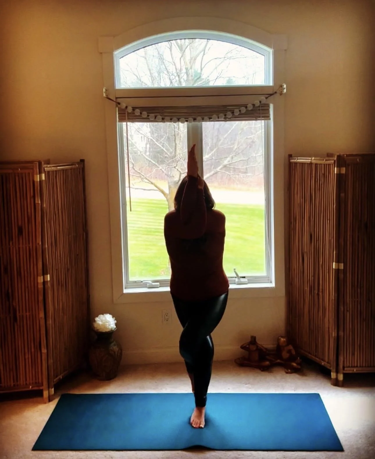 A woman standing on a yoga mat in a room, practicing yoga in front of a large window with a view of trees and a grassy yard outside.