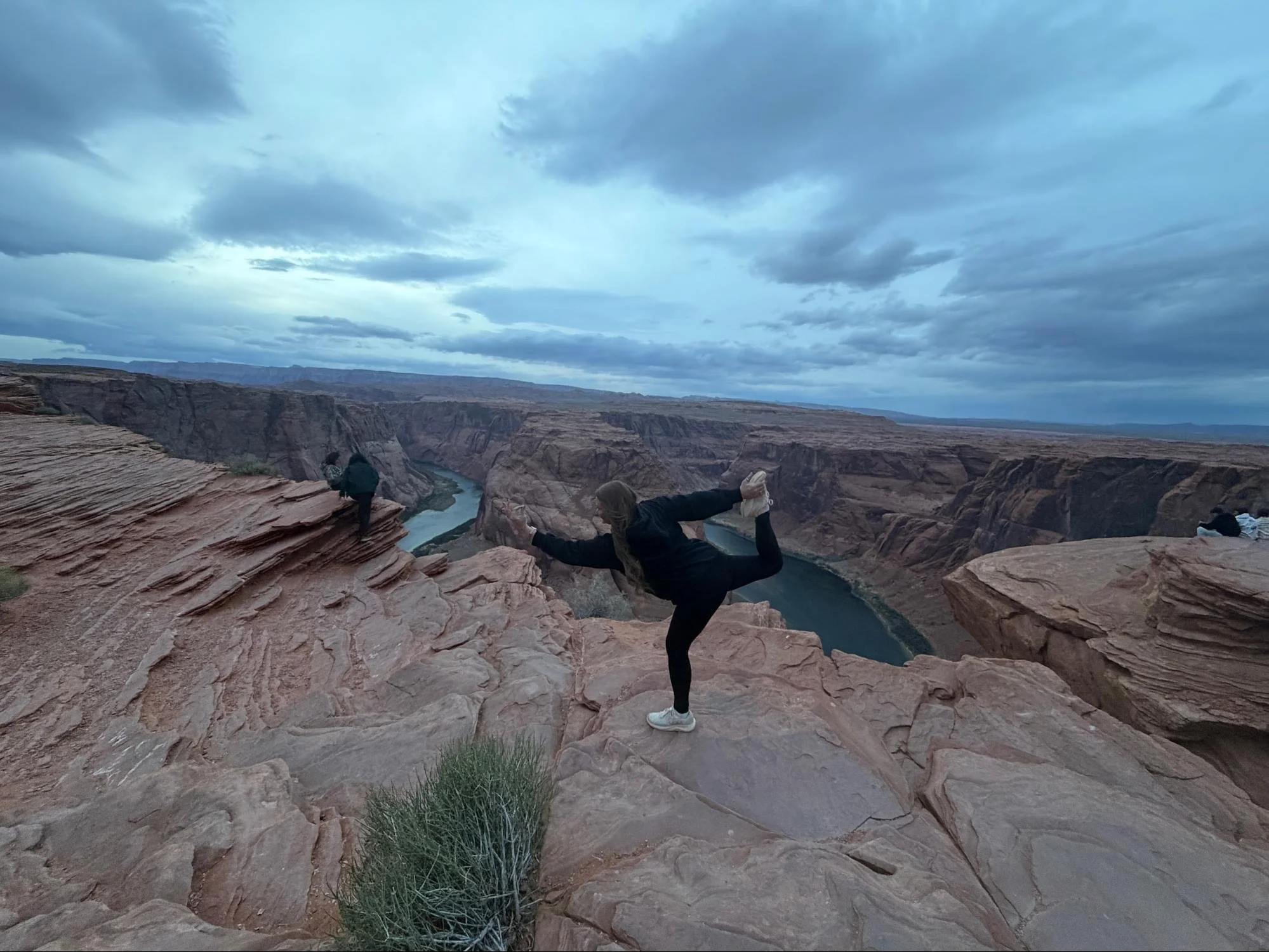 Two people are taking selfies on the edge of a canyon, with a river visible below and cloudy skies overhead.