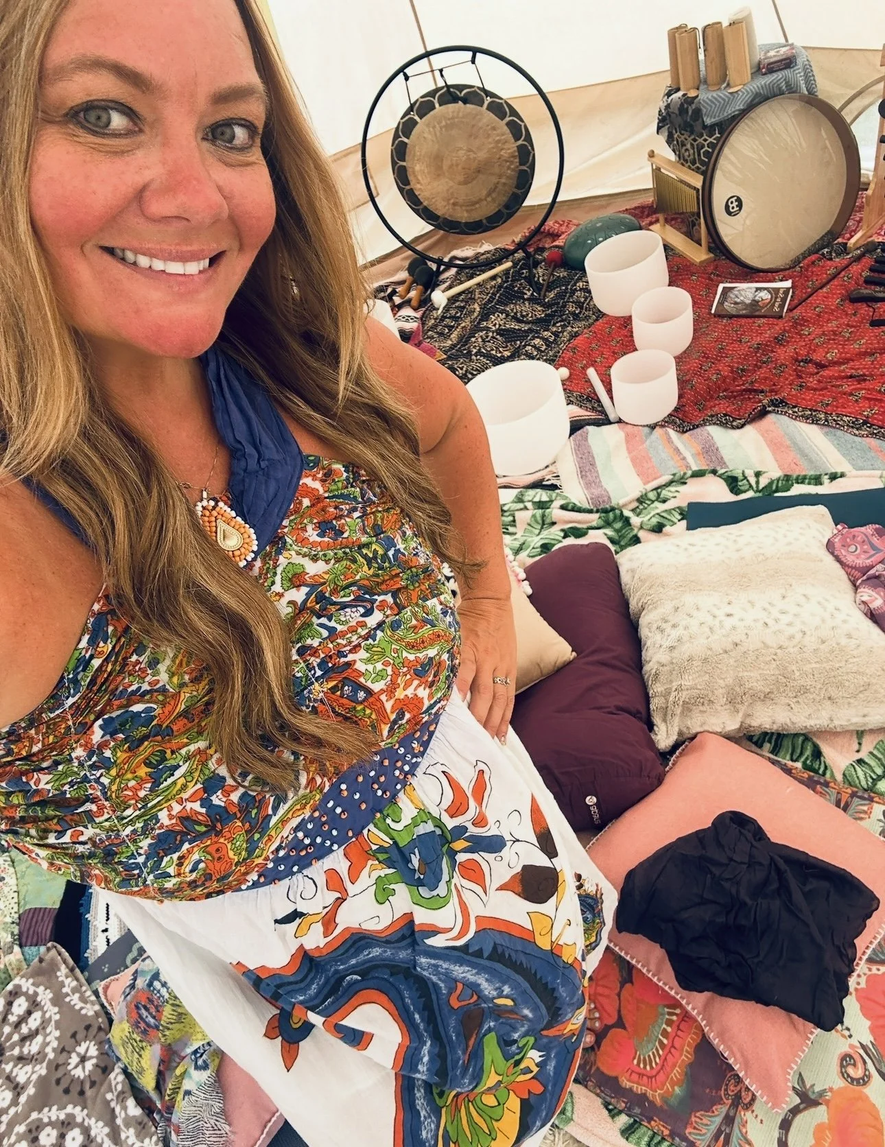 A woman with long, wavy hair smiling and wearing a colorful, patterned top and white apron, standing in front of a collection of musical instruments including singing bowls, a gong, and a drum, with various cushions and textiles in the background.