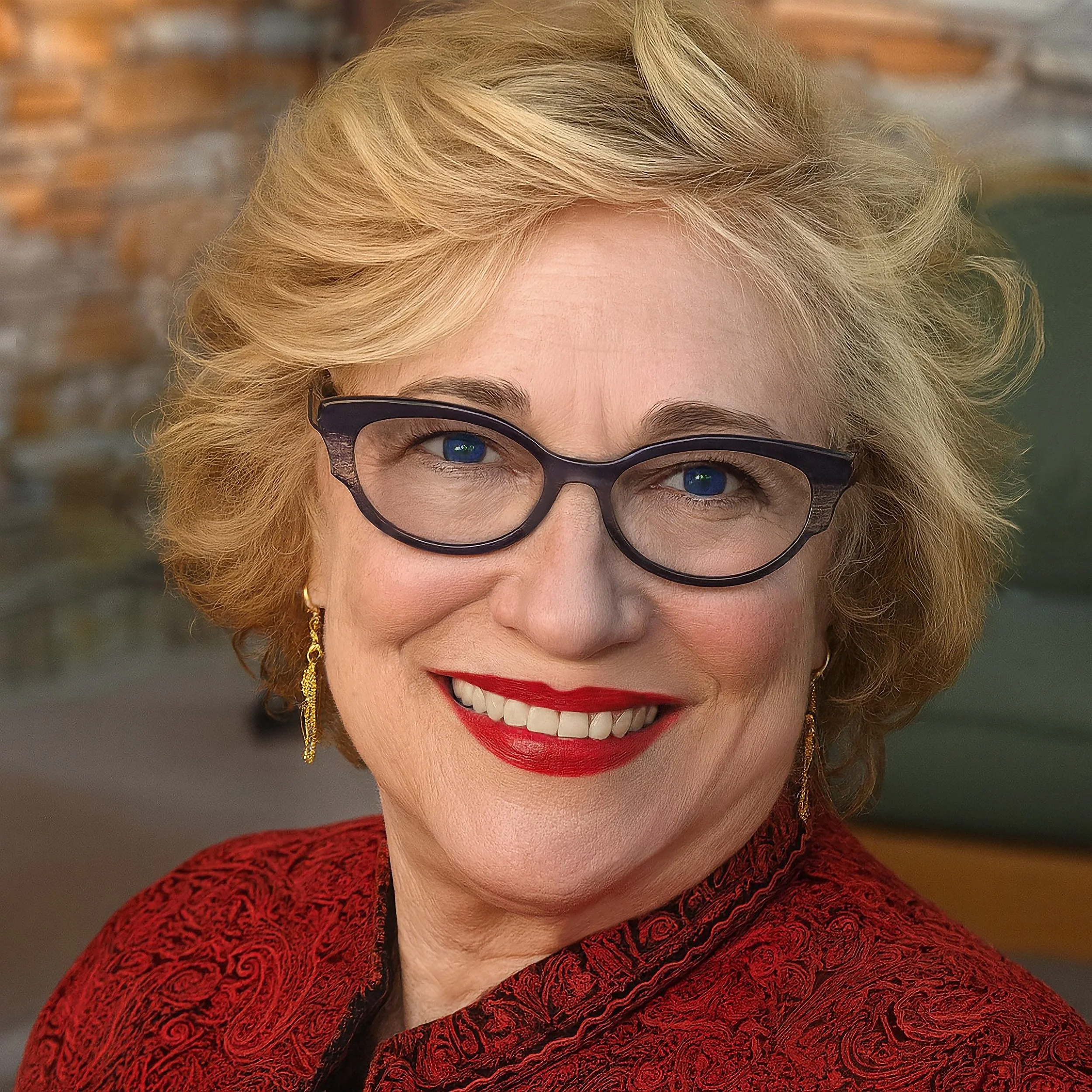 A smiling elderly woman with blonde hair, blue eyes, black-rimmed glasses, red lipstick, and earrings, wearing a red paisley-patterned blazer, in front of a blurred brick wall background.