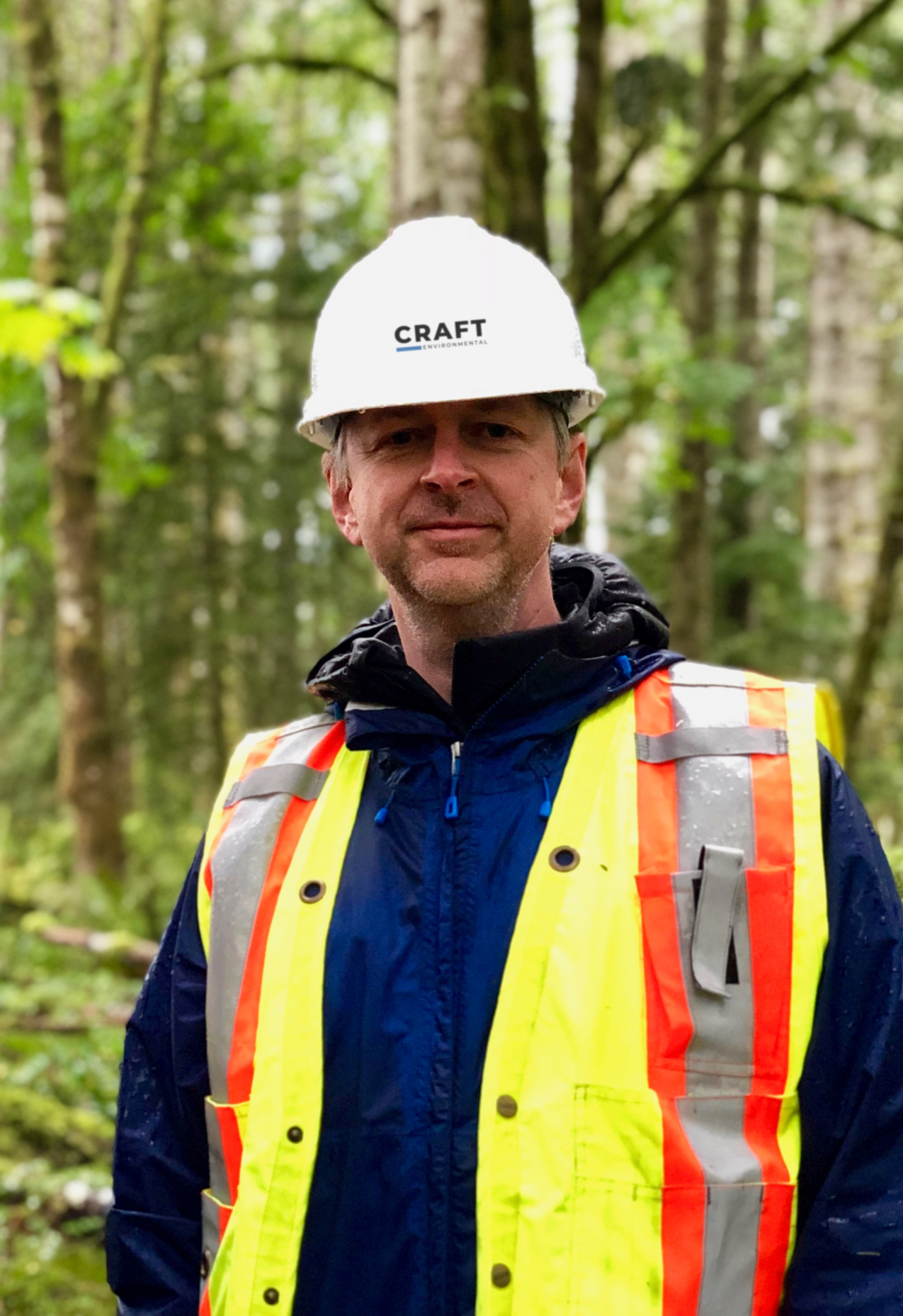 Geoff Wickstrom in a white hard hat and a high-visibility safety vest standing outdoors in a wooded area.