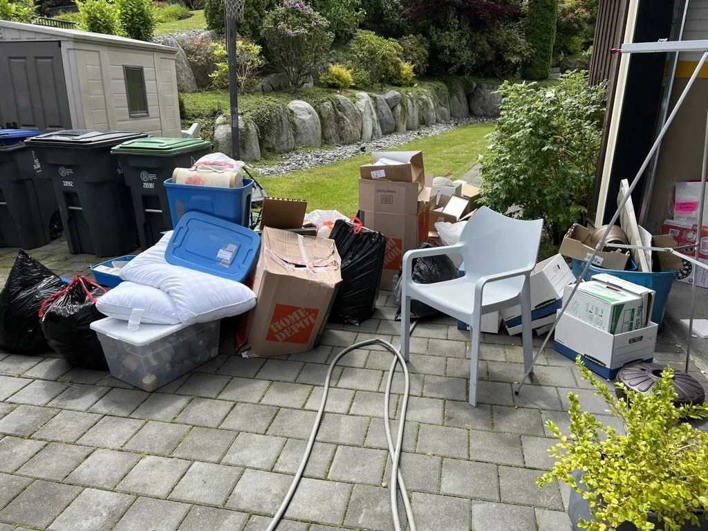 An outdoor patio with a pile of assorted boxes, trash bags, and household items including pillows, a plastic storage bin, and cardboard boxes. There's a white plastic chair, a garden hose, and a row of trash bins nearby. The background features a garden with rocks, grass, and bushes.