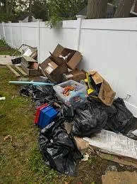 Pile of scattered trash and discarded boxes against a white fence in a yard.