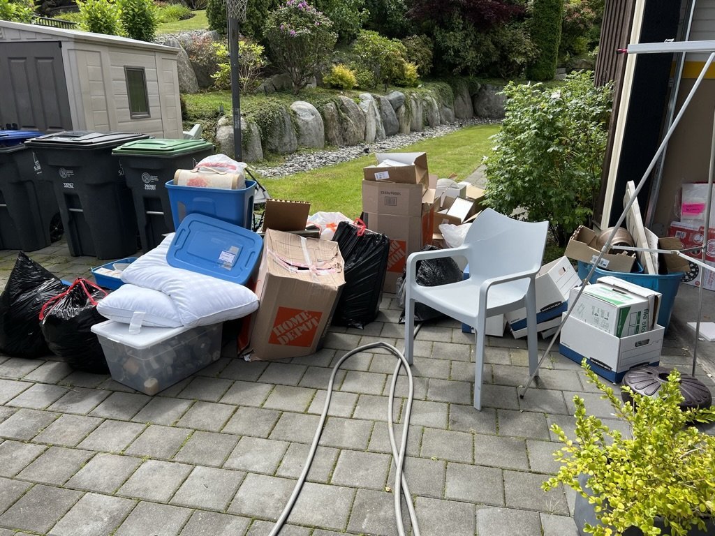 Cluttered outdoor patio with moving boxes, plastic containers, bags, chairs, and various household items, surrounded by a garden with rocks and greenery.