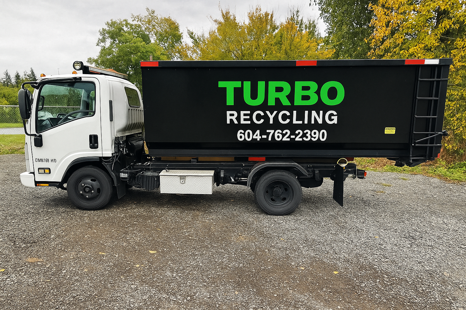 A white recycling truck with a black container on the back that has green and white text reading "TURBO RECYCLING" and a phone number, parked on a gravel lot with trees and a cloudy sky in the background.