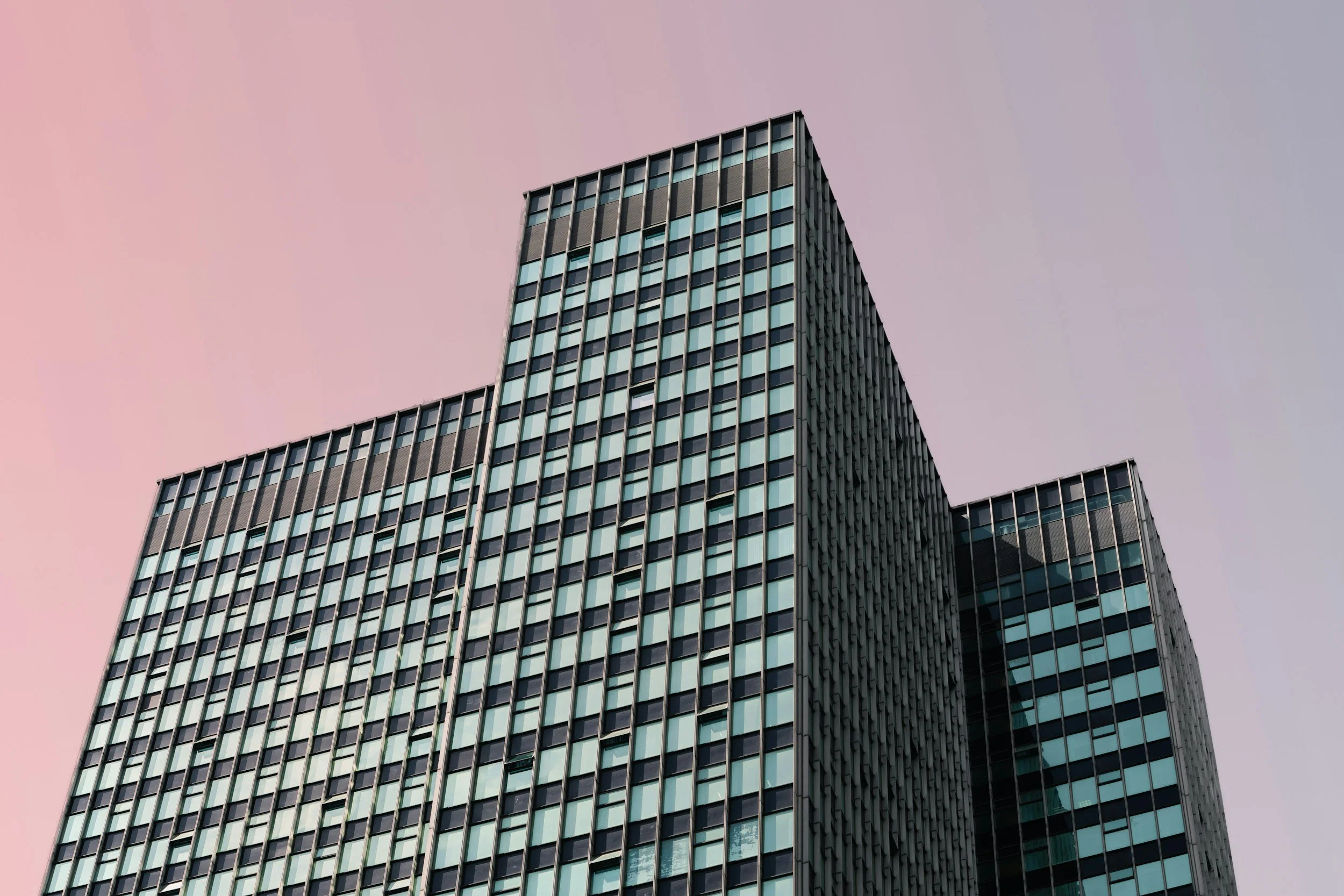 Modern high-rise office building with glass windows, set against a pastel pink and gray sky.