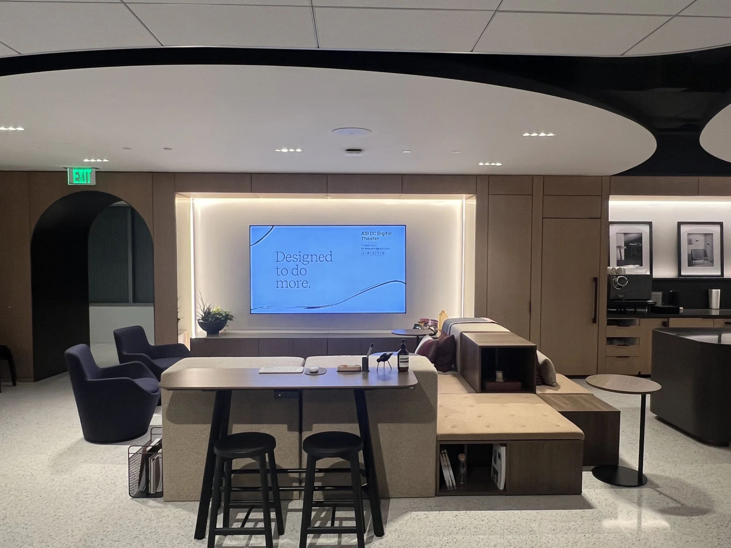 Modern office lounge with a large digital screen displaying the message 'Designed to do more.'; seating including armchairs and a sofa; a small side table; decorative pictures on the wall; ambient lighting.