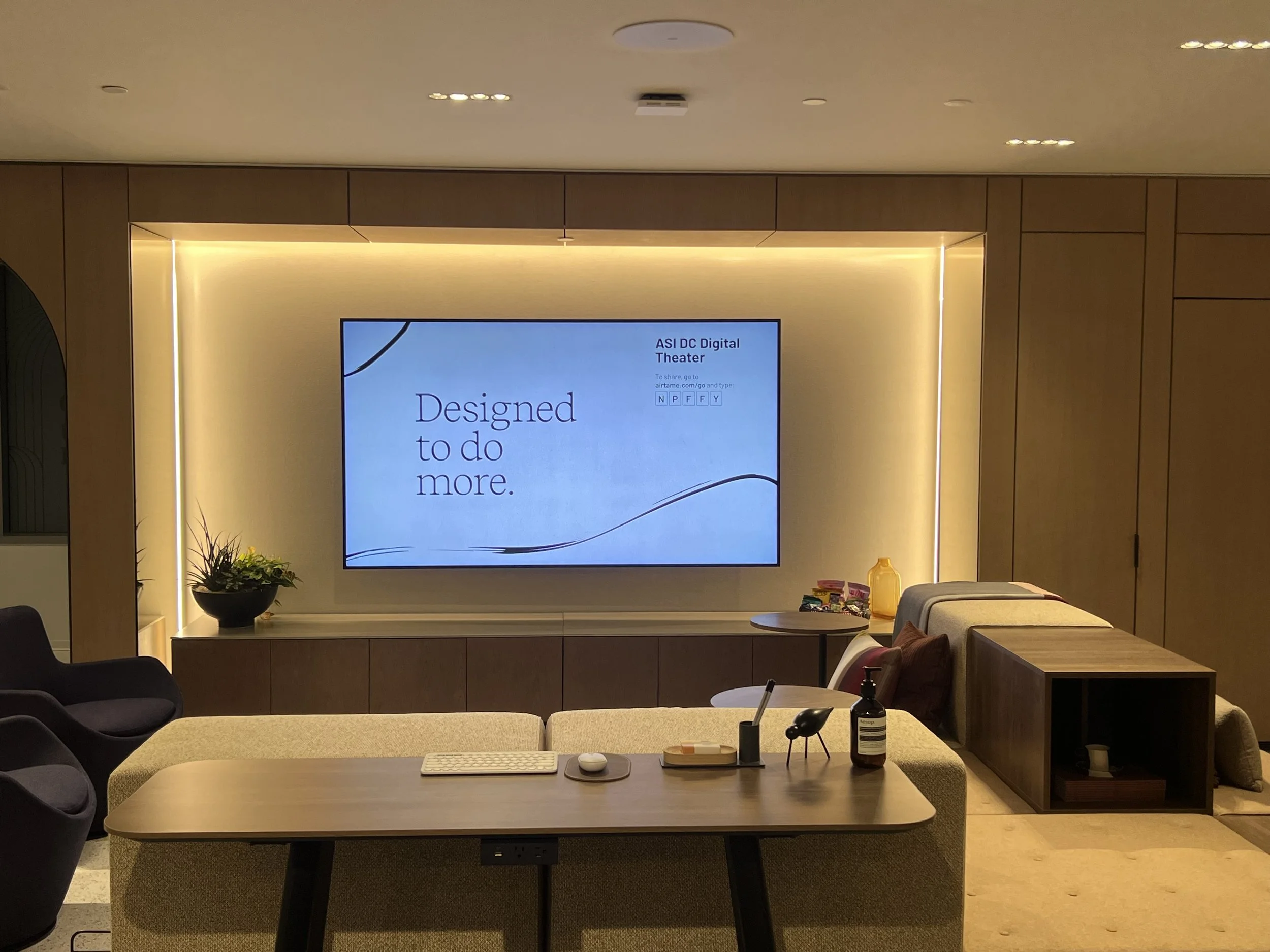 Interior of a modern lounge area with a large digital screen displaying the message 'Designed to do more.' The space features wooden wall panels, a beige couch, a dark armchair, and a small table with snacks and drinks.