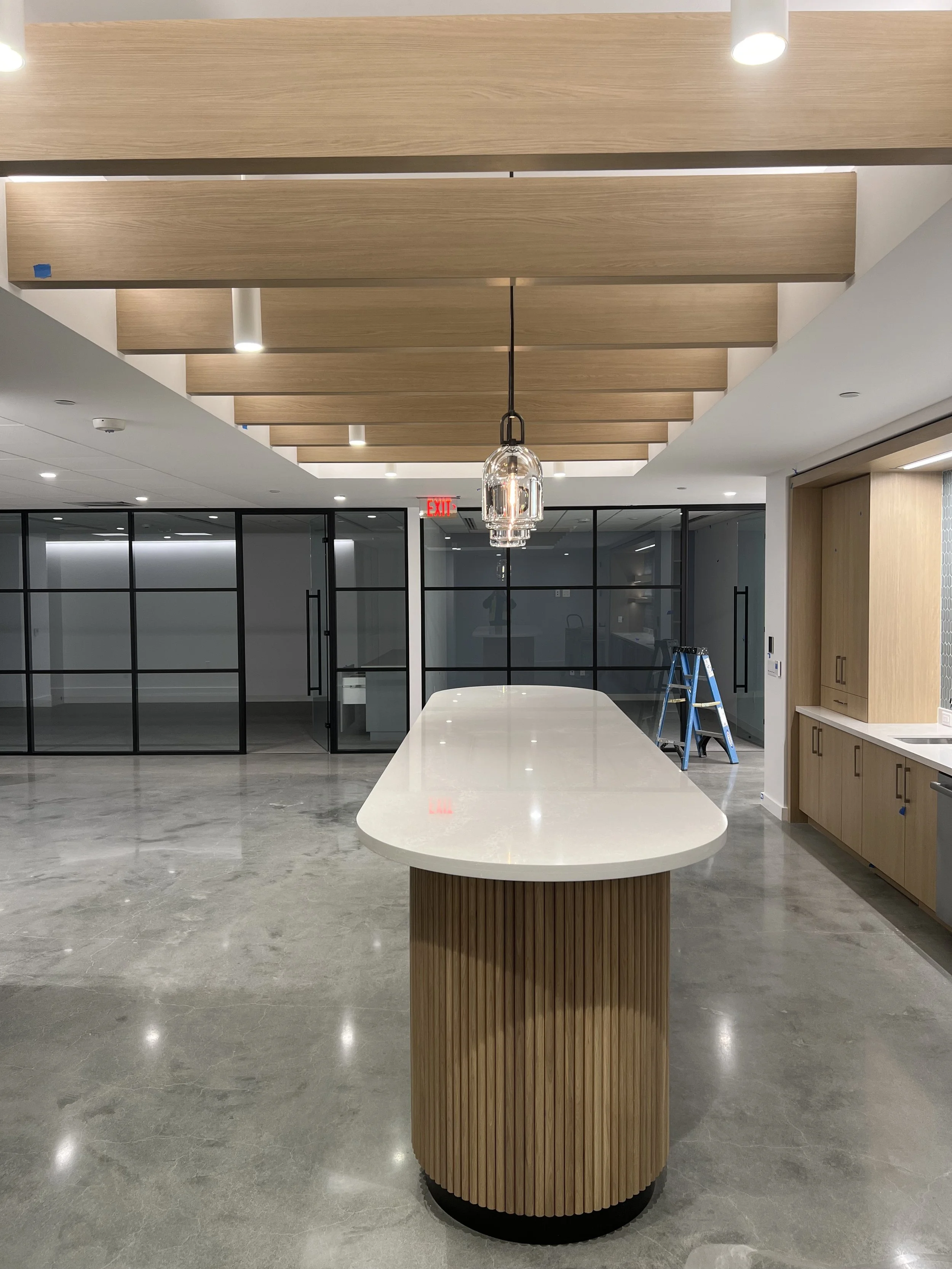 Modern kitchen or dining area with a large white oval table, wooden accents on the ceiling and table base, glass doors at the back, and a ladder on the side.