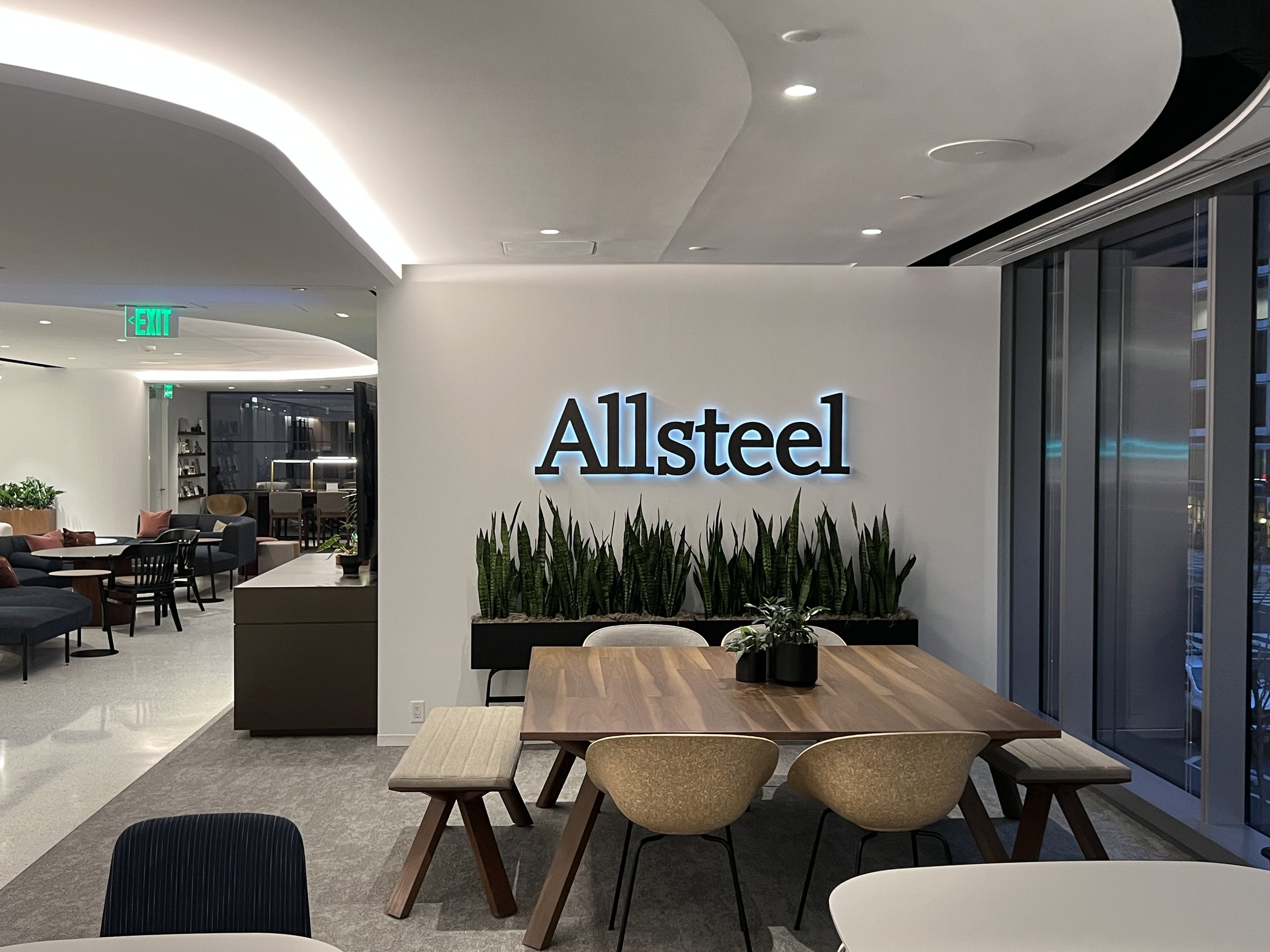 Modern office lobby with a sign that reads 'Allsteel' on the wall, a wooden table with three chairs, and a planter with tall green plants. In the background, there are seating areas with chairs and tables, and large windows with blinds.