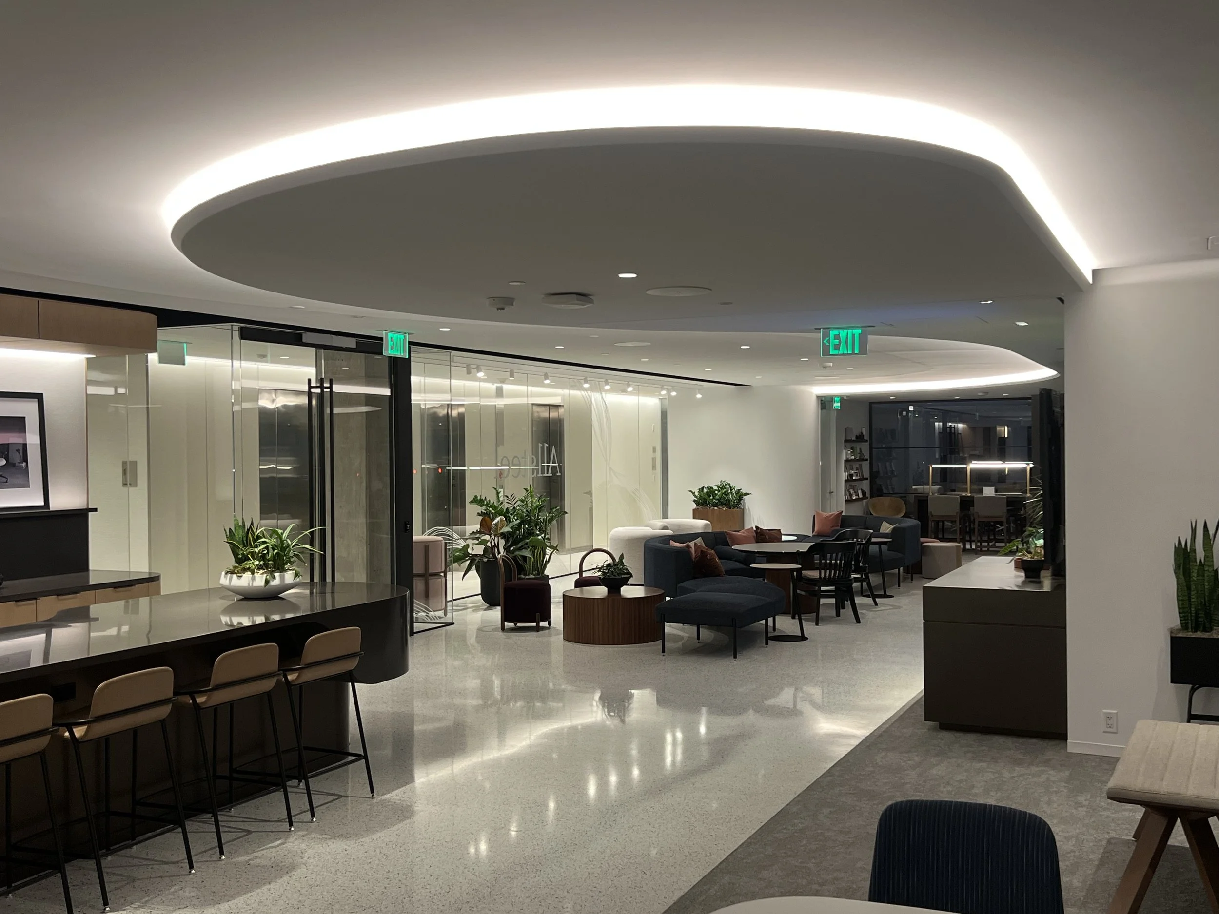 Modern lounge area with armchairs, sofas, tables, and potted plants, illuminated by soft overhead lighting. Glass walls and exit signs are visible.