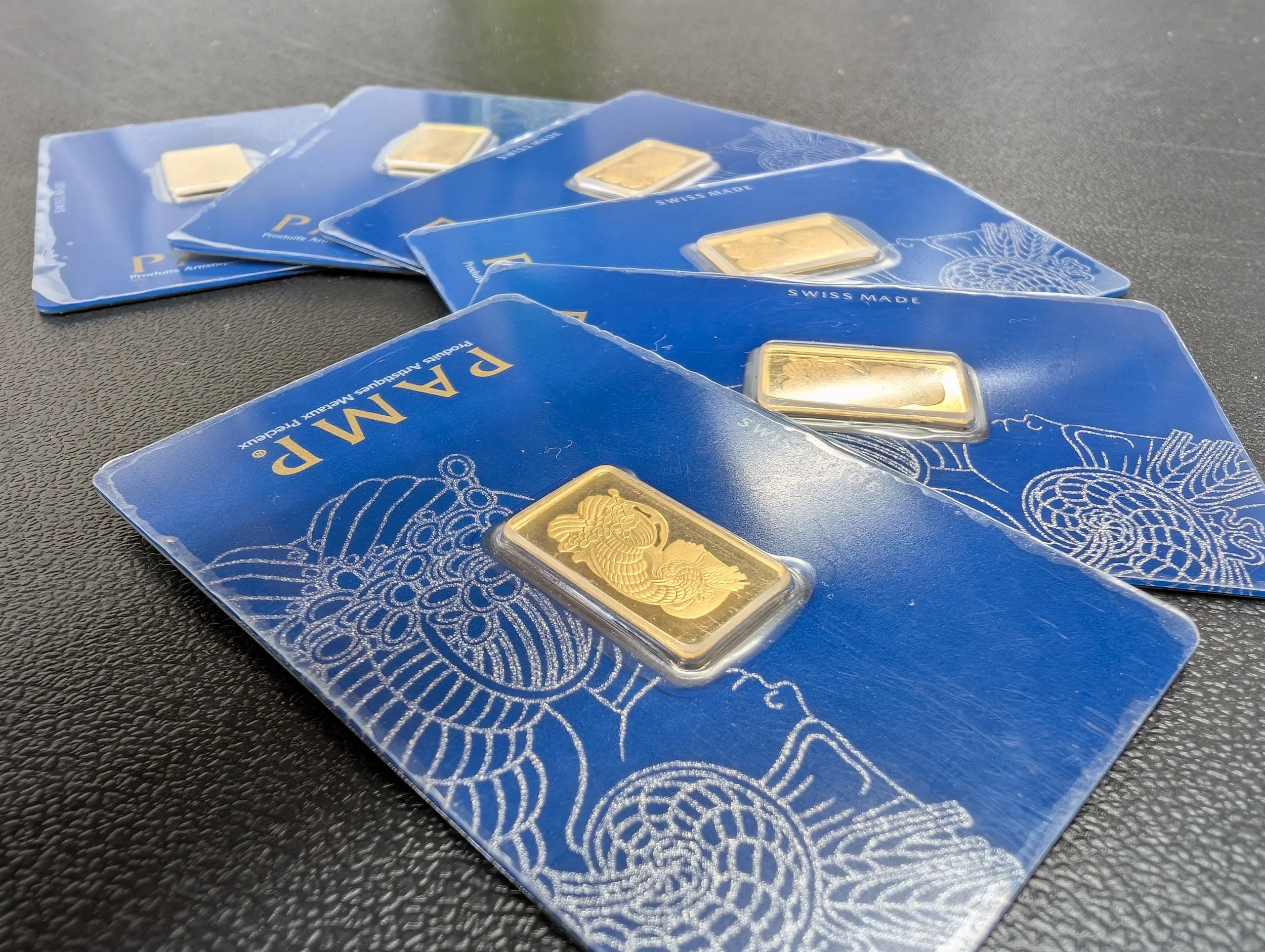 Multiple blue packages of 'PAM P.' gold bars with dragon designs, spread out on a dark textured surface.