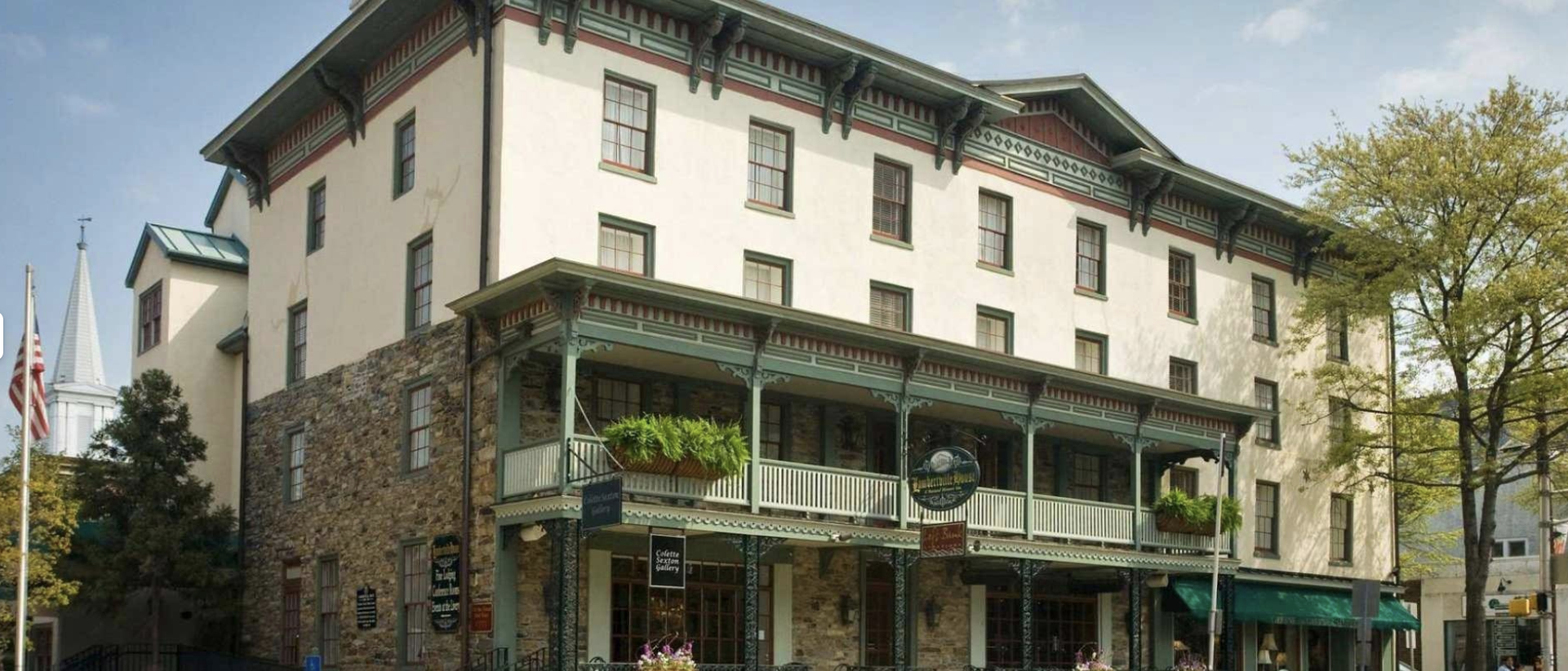 Historic four-story building with a stone ground floor and a light-colored upper floors, featuring a large balcony with green railings and hanging flower baskets, situated on a street with trees and painted flags.