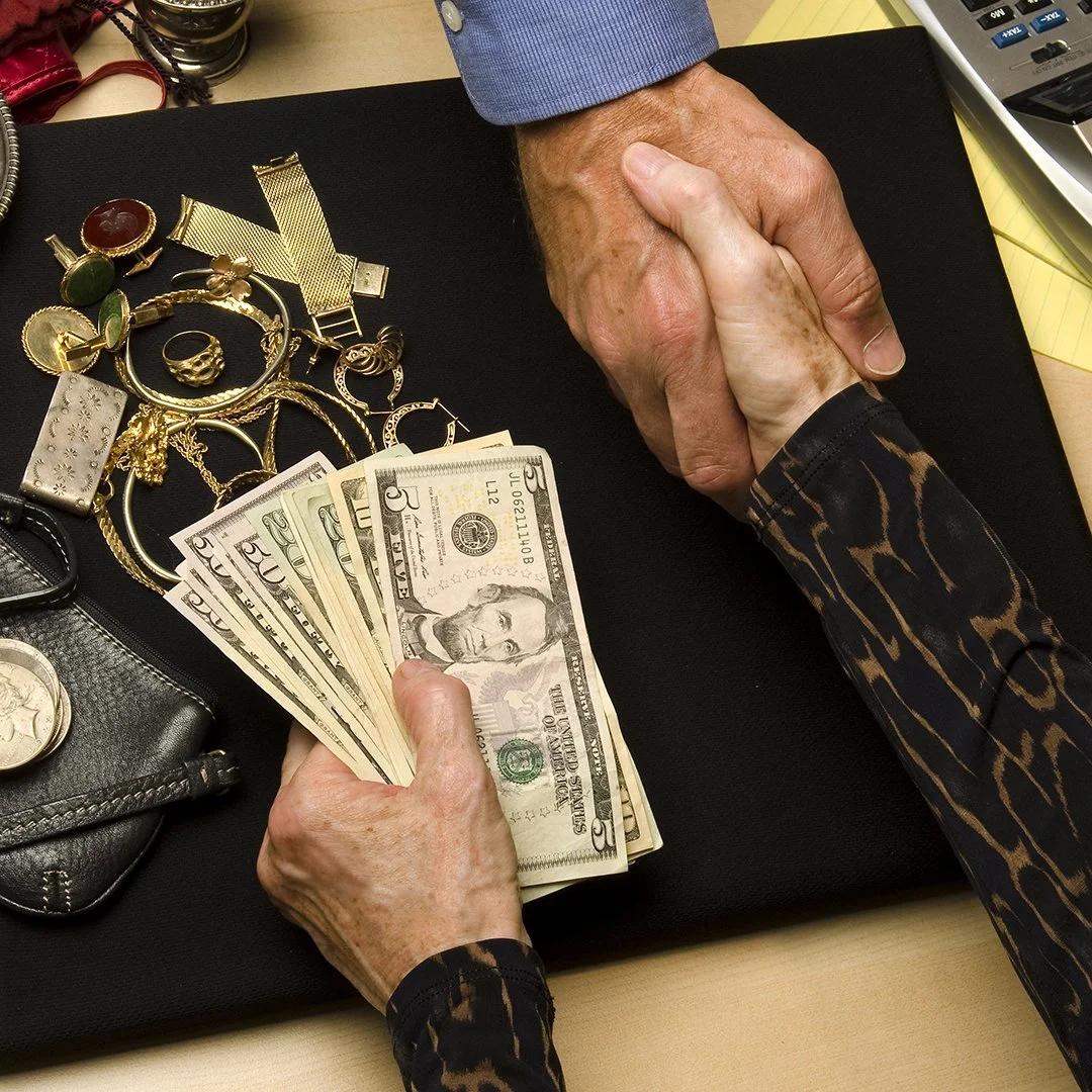 An elderly person and a younger person shaking hands over a table with cash and jewelry laid out, including a five-dollar bill, gold rings, watches, and necklaces.