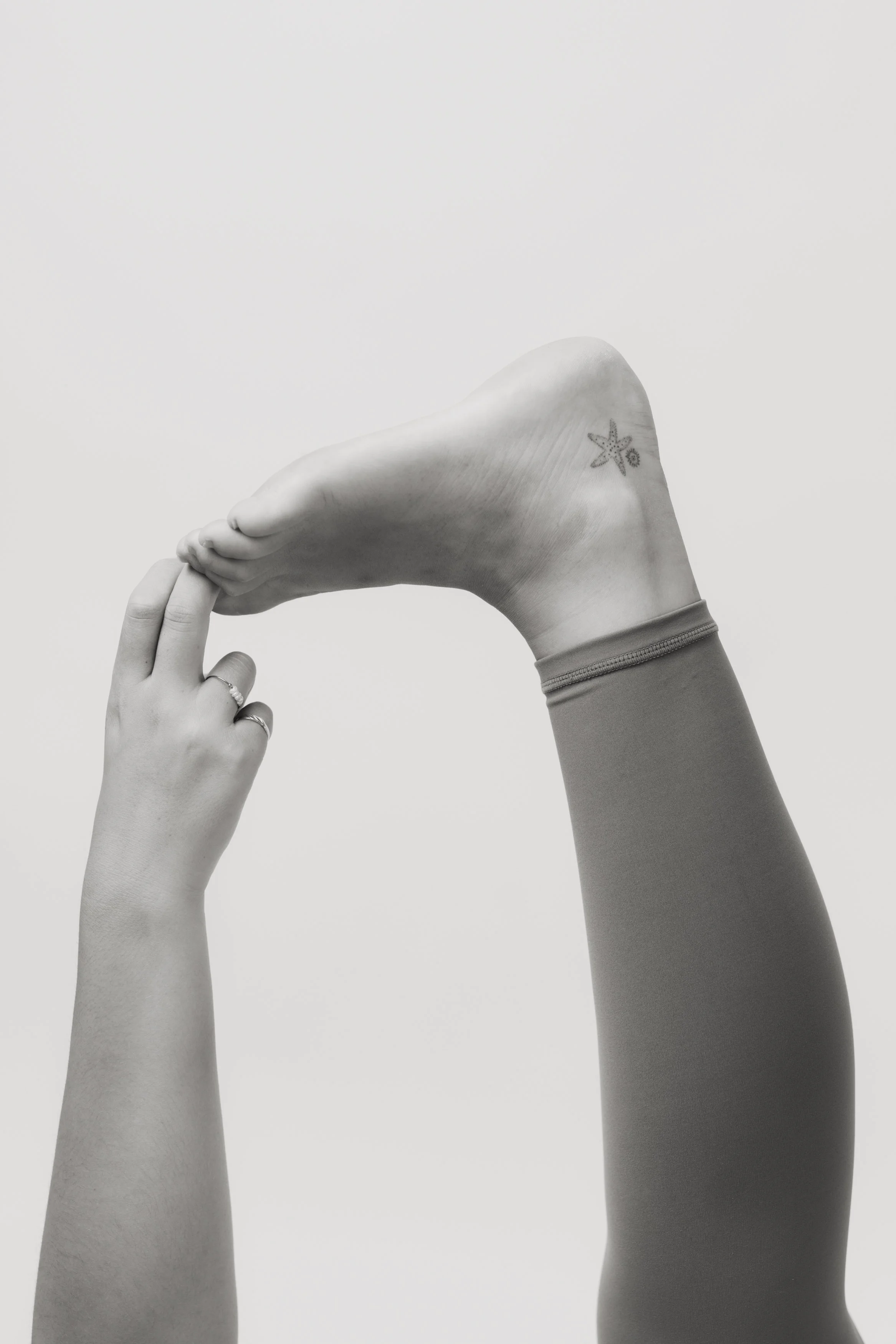 A person with a tattoo on their ankle, shows their foot, with one hand holding their toes, against a plain background.