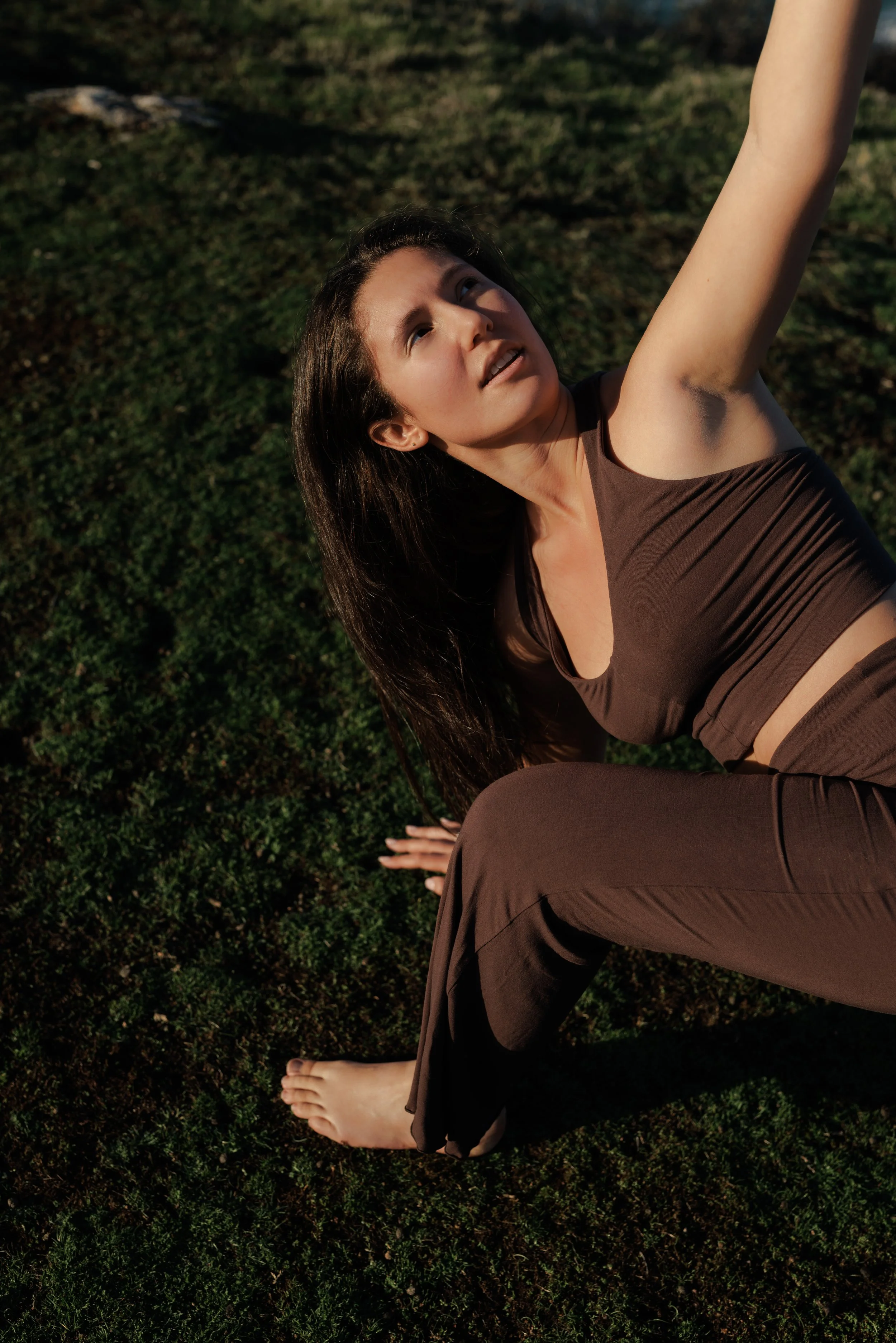 A woman with long dark hair in brown clothing practicing yoga outdoors on grass, in a pose with one arm extended upward and the other hand on the ground.