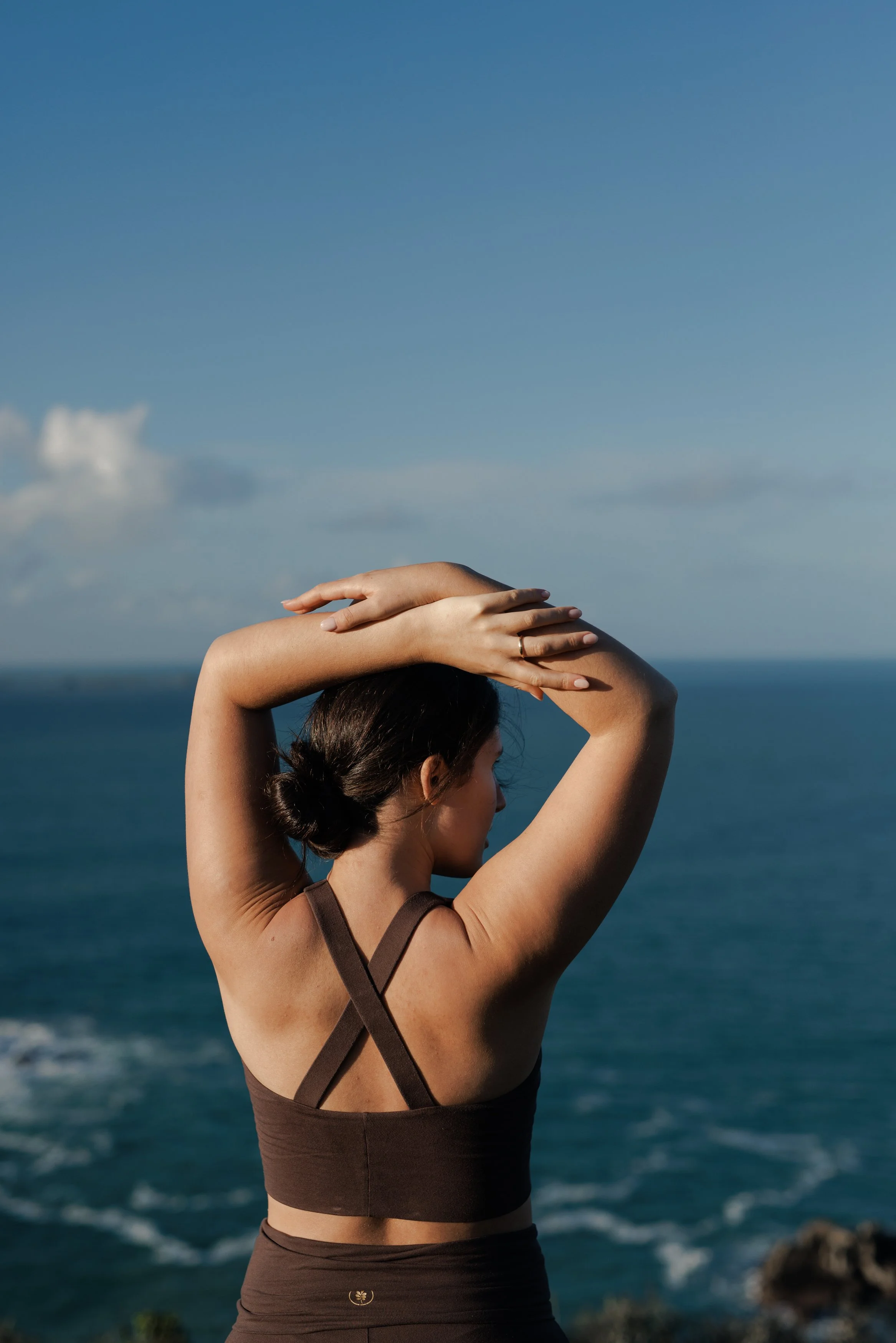 A woman in a sports bra standing outdoors with her back to the camera, arms raised and hands resting on her head, overlooking the ocean with a clear blue sky.