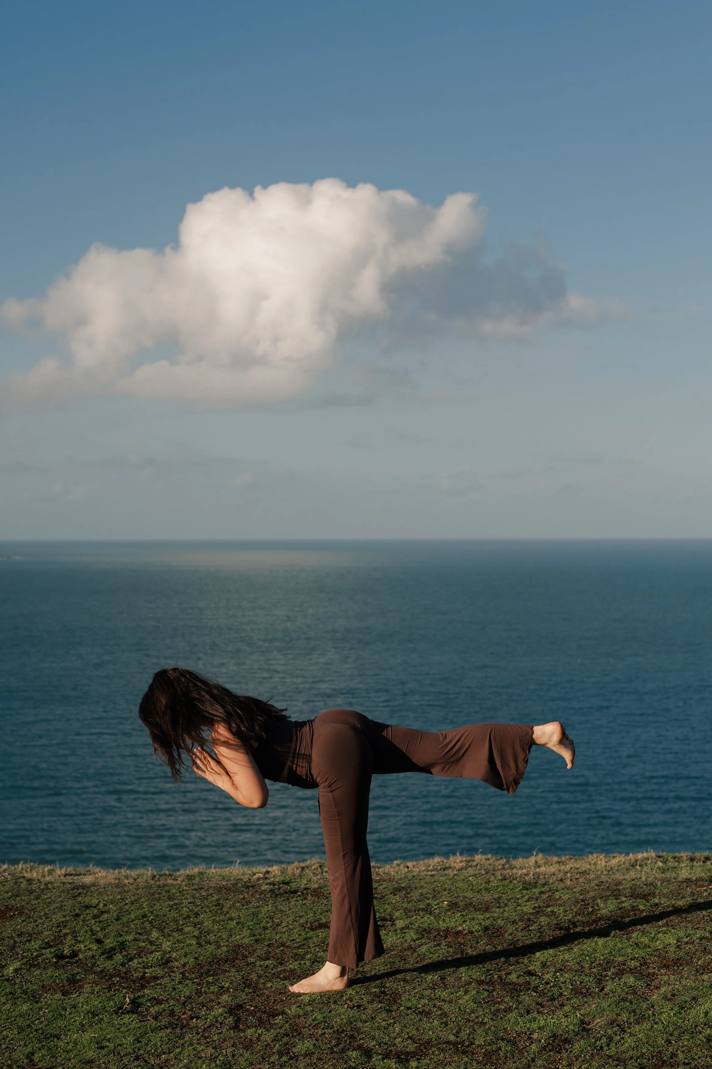 Person practicing yoga outdoors on grass near water, performing a balance pose with one leg raised and hands covering face, ocean and sky in background.