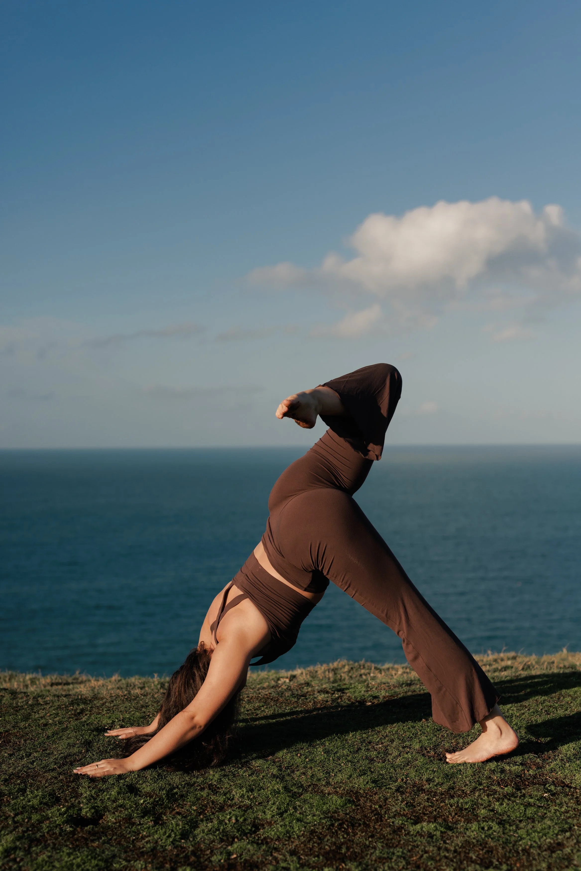 Woman practicing yoga outdoors on grass, performing downward dog pose with ocean and sky in background.