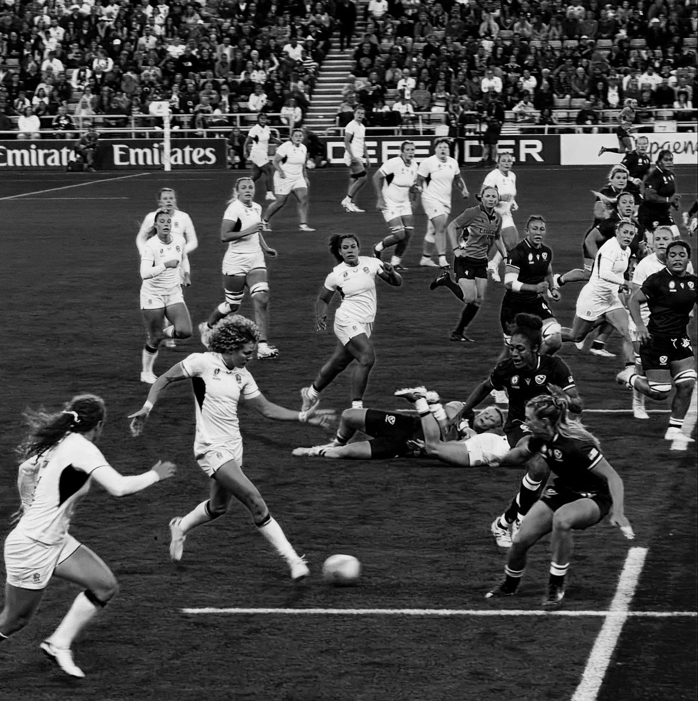 Women soccer game, players running and chasing the ball on the field, some players in white jerseys, some in dark jerseys, a crowd of spectators in the stands, black and white photo.