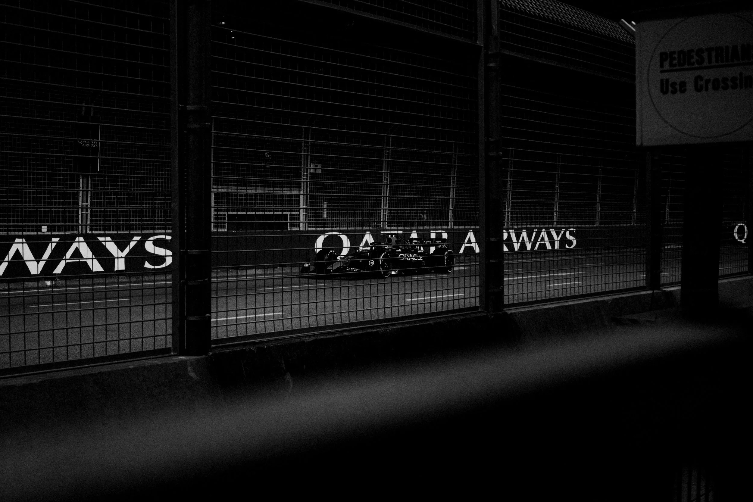 Black and white photograph of a Formula 1 race car speeding on a race track, viewed through a metal fence with signage that reads 'QATAR AIRWAYS' and 'PEDESTRIAN Use Crosswalk'.