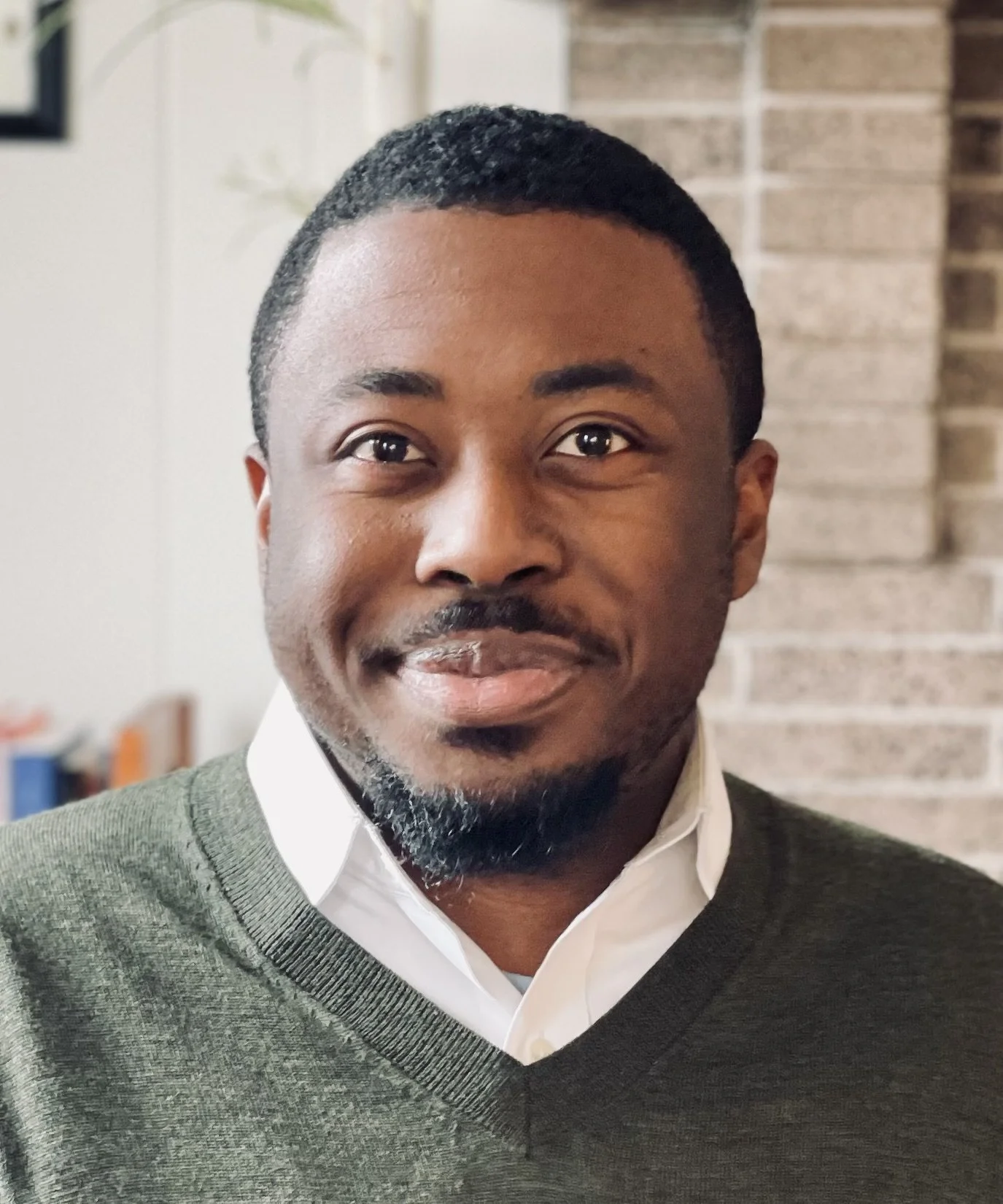 A man with short black hair, a goatee, wearing a white shirt and green sweater, smiling, with a brick wall in the background.