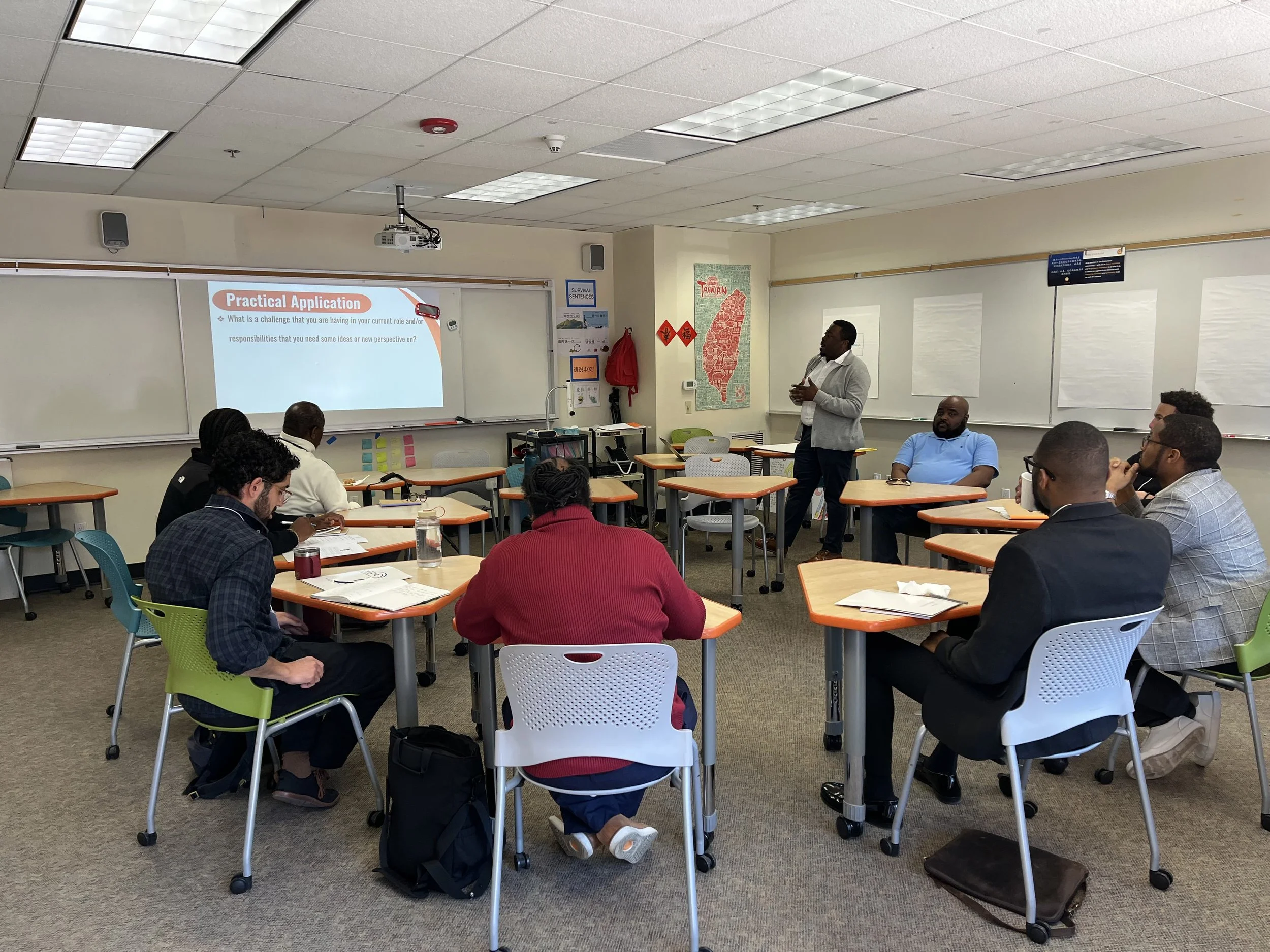 A group of people attending a presentation in a classroom with a whiteboard. The presenter is standing near the whiteboard, which displays a slide titled 'Practical Application'.