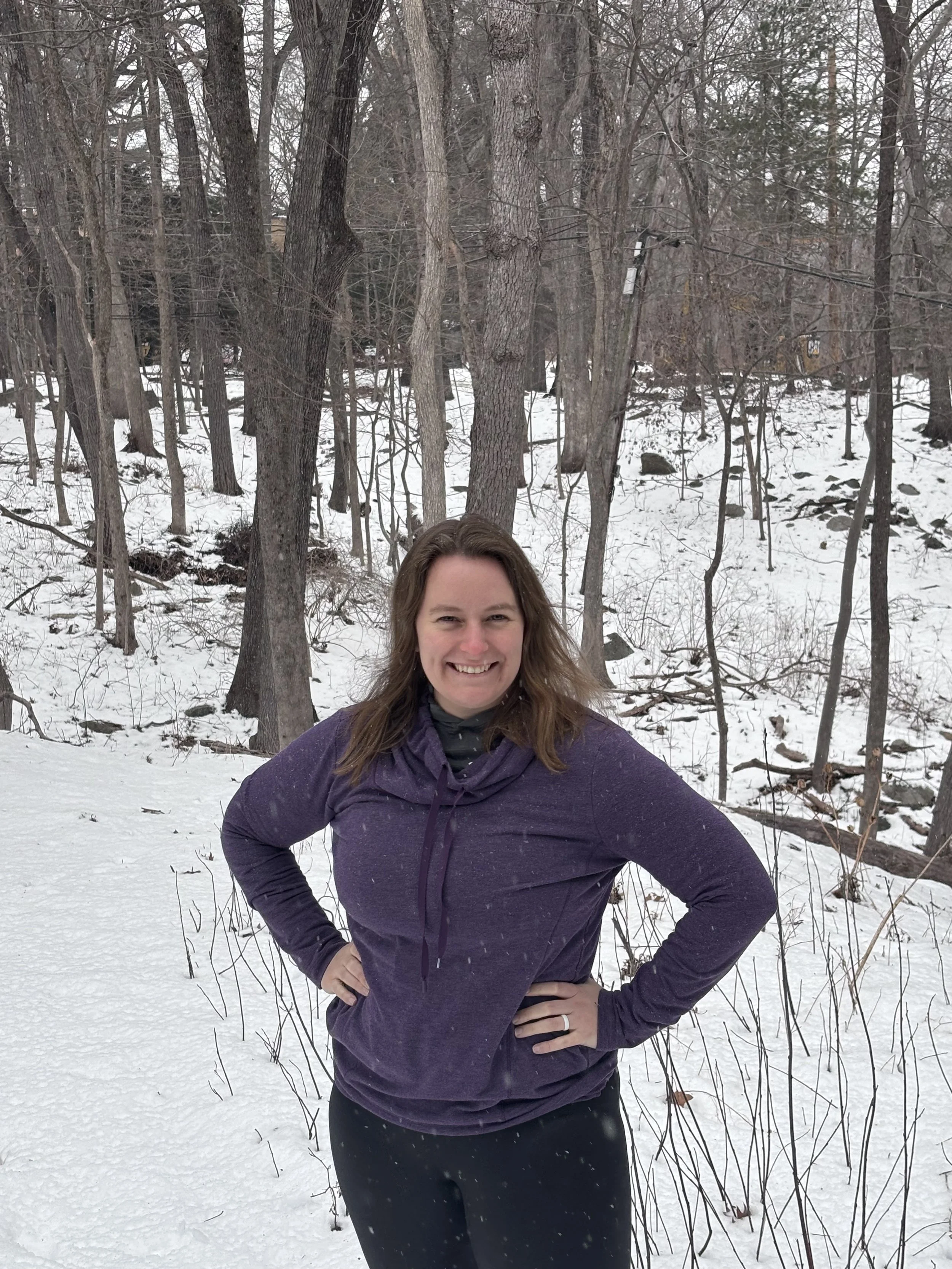 A woman smiling outdoors in a snowy forest, wearing a purple jacket and black pants.
