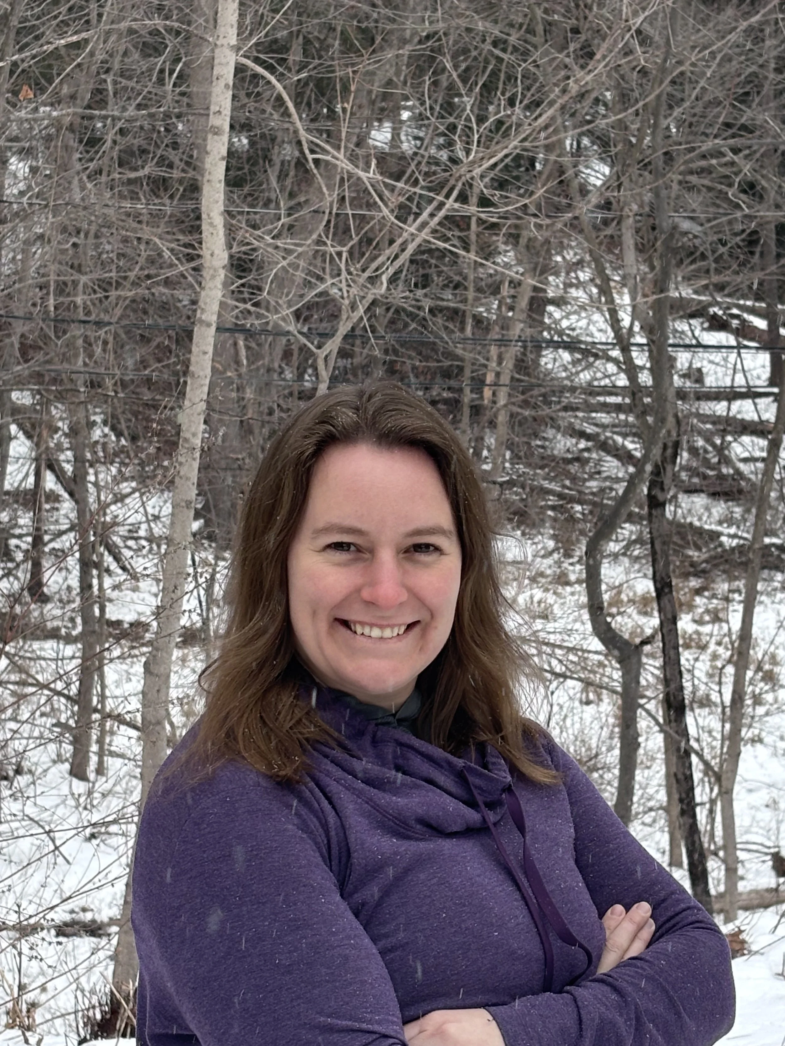 A woman with shoulder-length brown hair smiling outdoors in a snowy forest, wearing a purple jacket with her arms crossed.