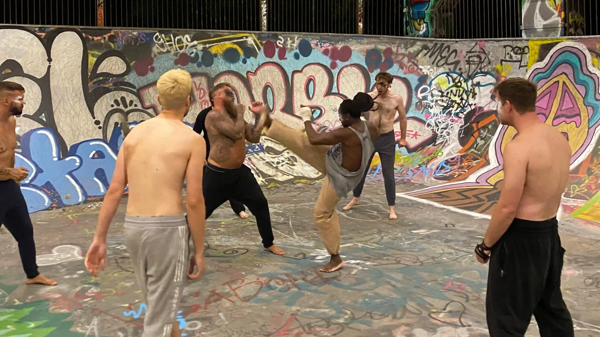 Two people set up a scene outdoors for filming or photography, with one adjusting a camera on a tripod and the other looking through it. There are objects on a wooden table, including a jar with a straw, a tumbler, and crumpled tissues. The background shows a grassy area and part of a house.