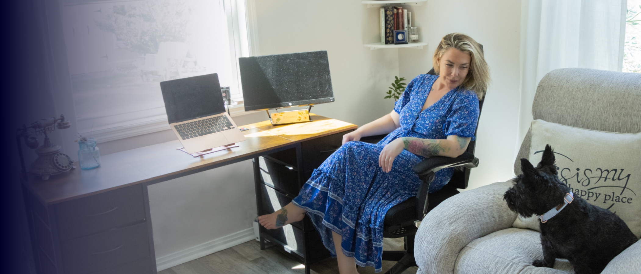 Woman sitting in a home office with a black dog on a couch nearby. She is wearing a blue dress with floral pattern, sitting in an office chair, and looking down. The desk has a laptop, two monitors, and a tissue box. There is a window behind the desk with sunlight coming through, and a white wall with a shelf holding books and decor.