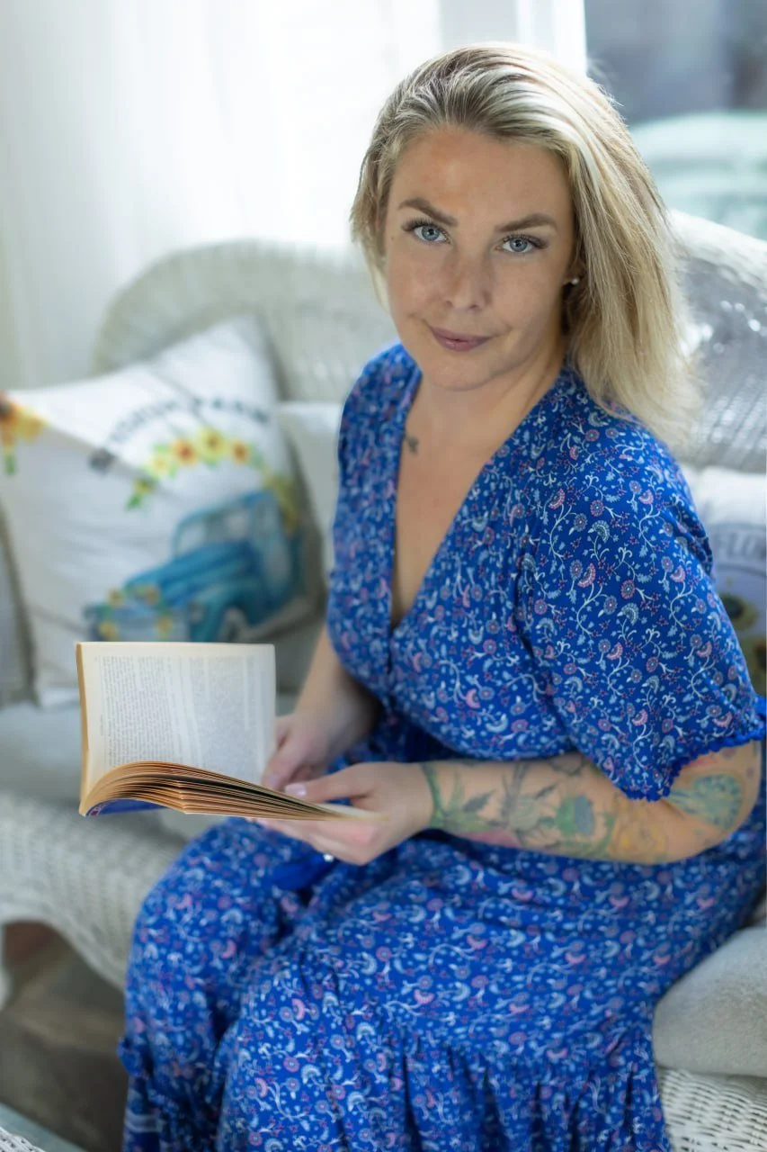 A woman with blonde hair and blue eyes sitting on a wicker sofa inside, holding an open book, wearing a blue floral dress, with tattoos on her left arm, and looking at the camera.