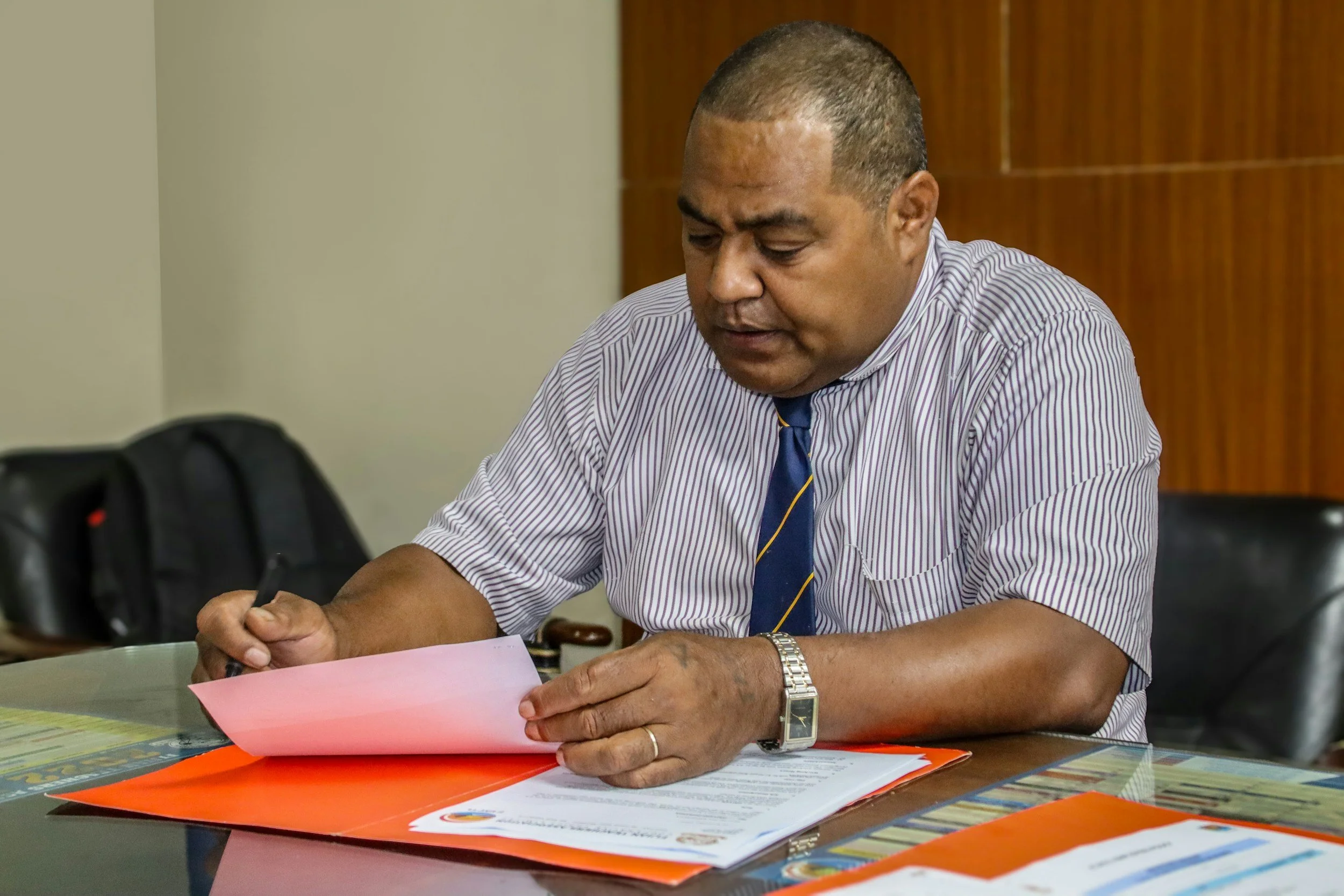 A man in a striped shirt and tie sitting at a table, reading and reviewing documents.
