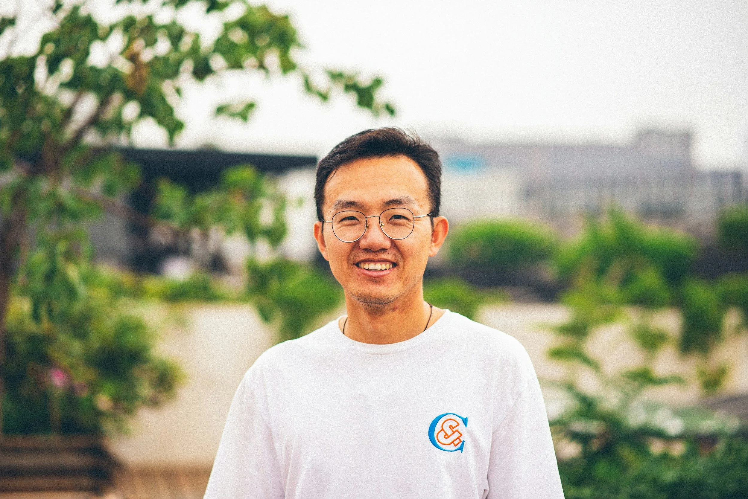 A smiling man wearing glasses and a white t-shirt with a logo, standing outdoors with green trees and buildings in the background.