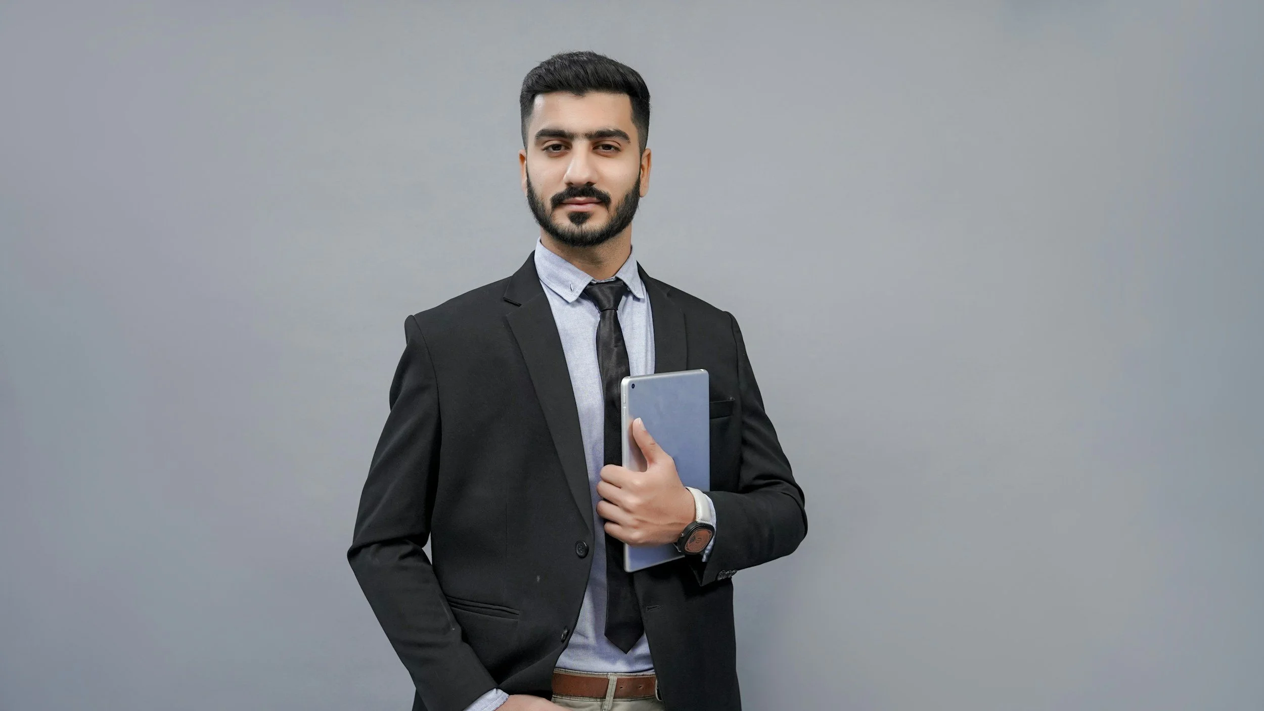 A young man in business attire, including a black suit, shirt, and tie, holding a tablet and standing against a plain gray background.