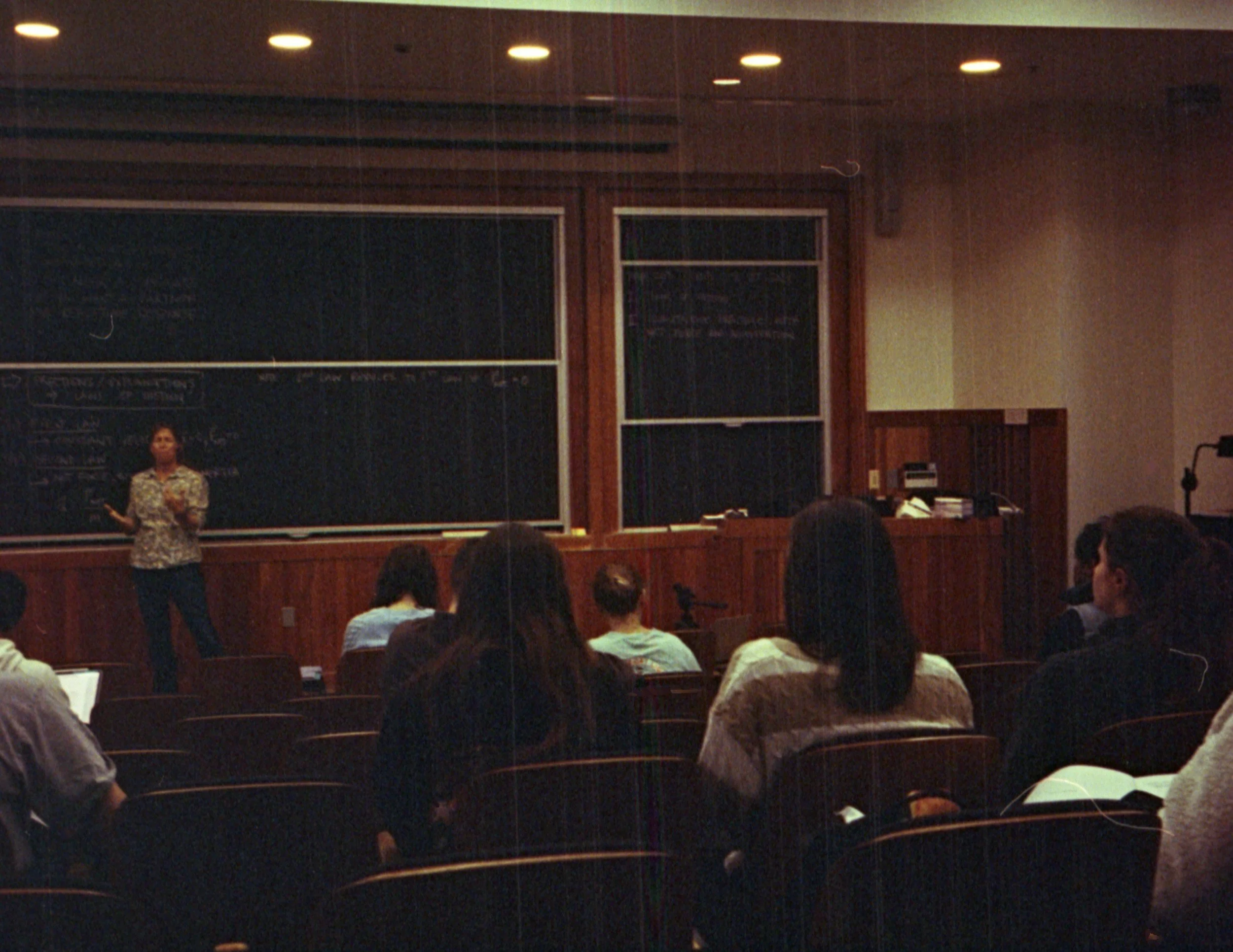 A woman stands in front of a chalkboard and presents to a classroom of students in a lecture hall with wooden walls and ceiling lights.