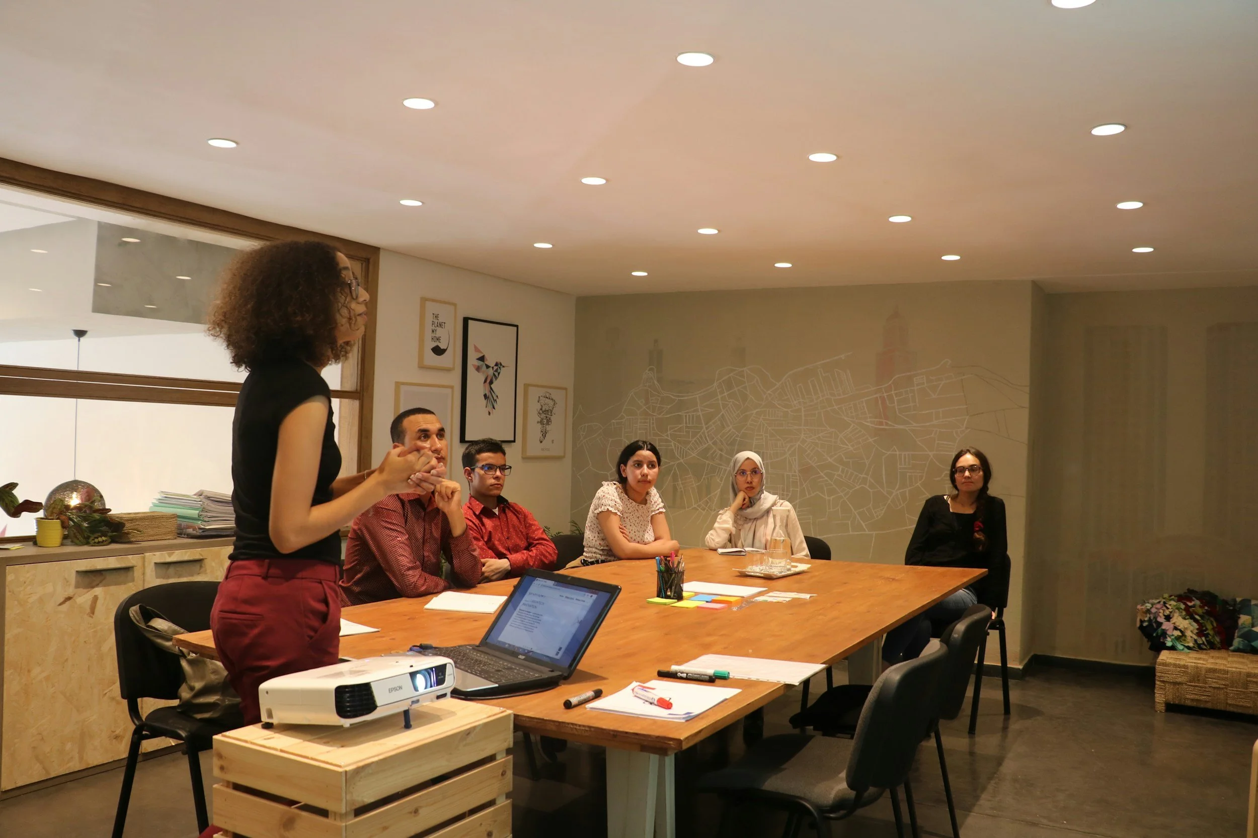A woman standing and speaking to a seated group of five diverse individuals in a modern conference room, with a laptop and projector on the table.