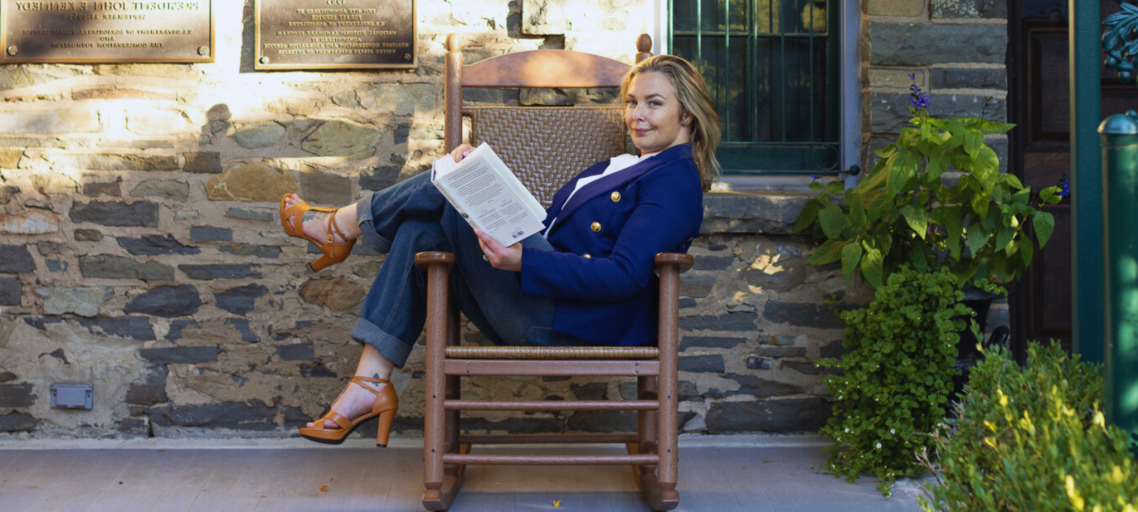 Woman with blonde hair sitting on a wooden chair outside, reading a book, beside a stone building with plants and plaque on the wall.