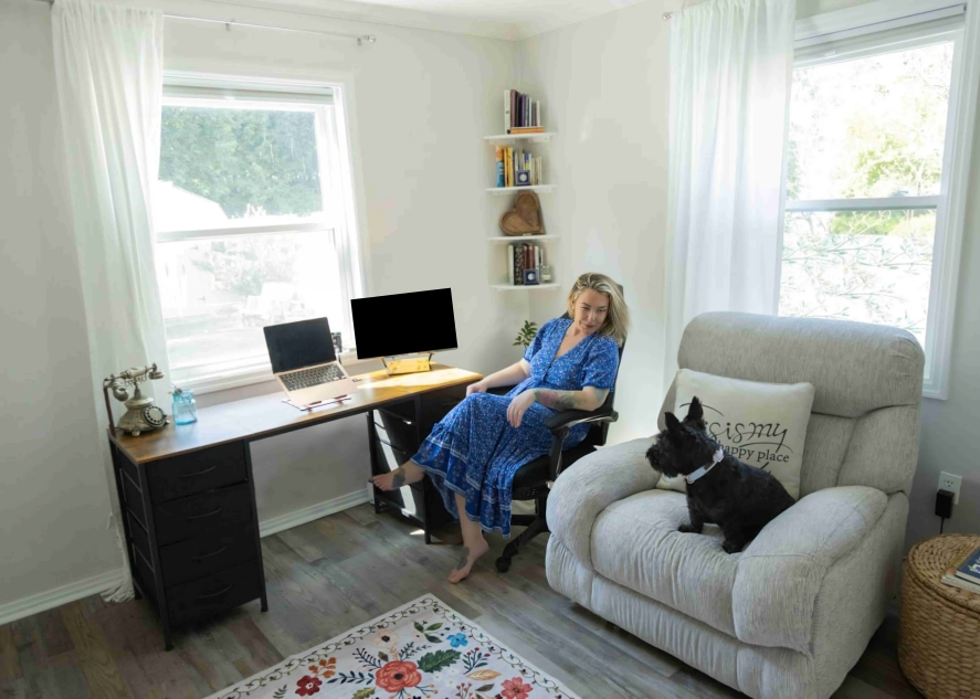 A woman in a blue dress sitting at a desk with two computer monitors and a laptop, next to a large beige armchair with a black dog on it, in a bright home office with white walls and large windows.