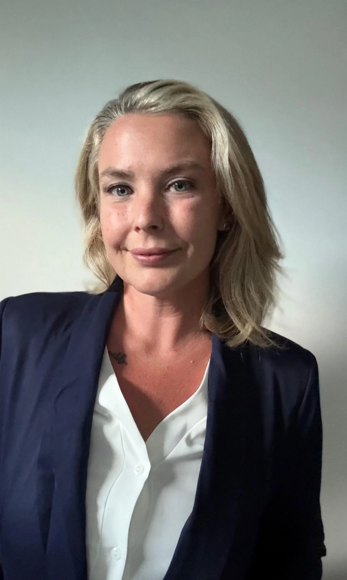 A woman with blonde hair wearing a white blouse and navy blazer is smiling at the camera against a light-colored wall.