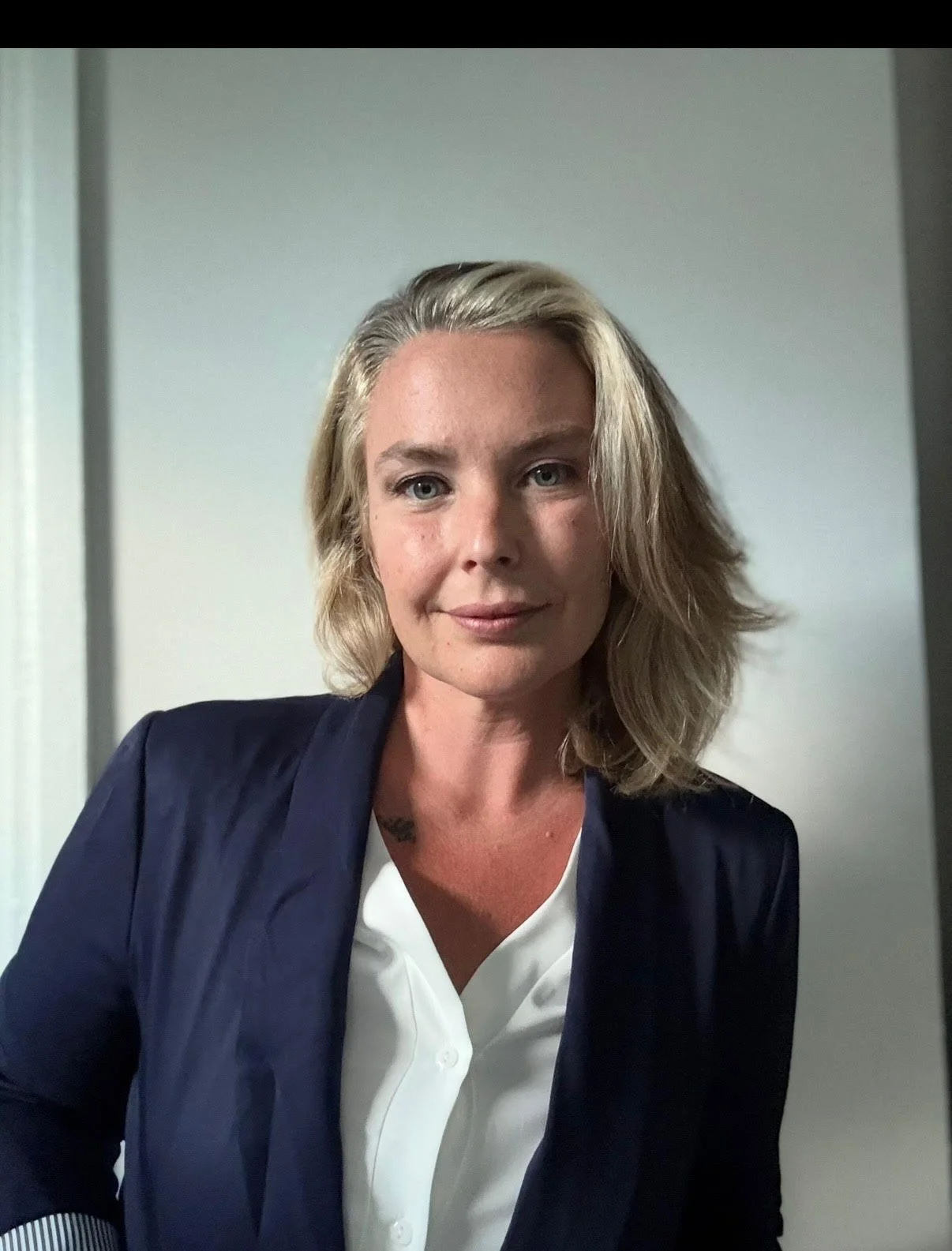 A woman with blonde hair, wearing a navy blazer and white blouse, sitting indoors against a light gray wall.