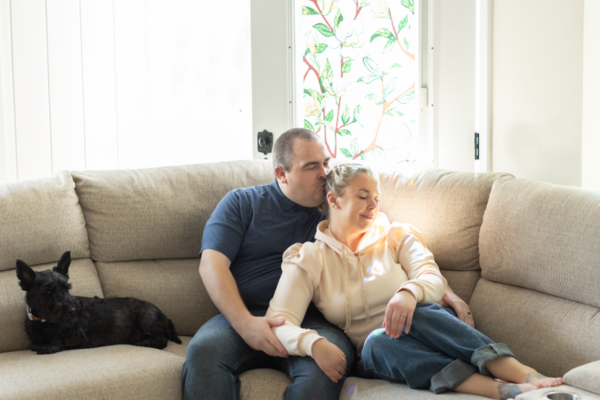 A couple sitting on a beige sofa, with the man kissing the woman on the head, and a black dog lying beside them. Behind them is a window with colorful plant-patterned curtains.