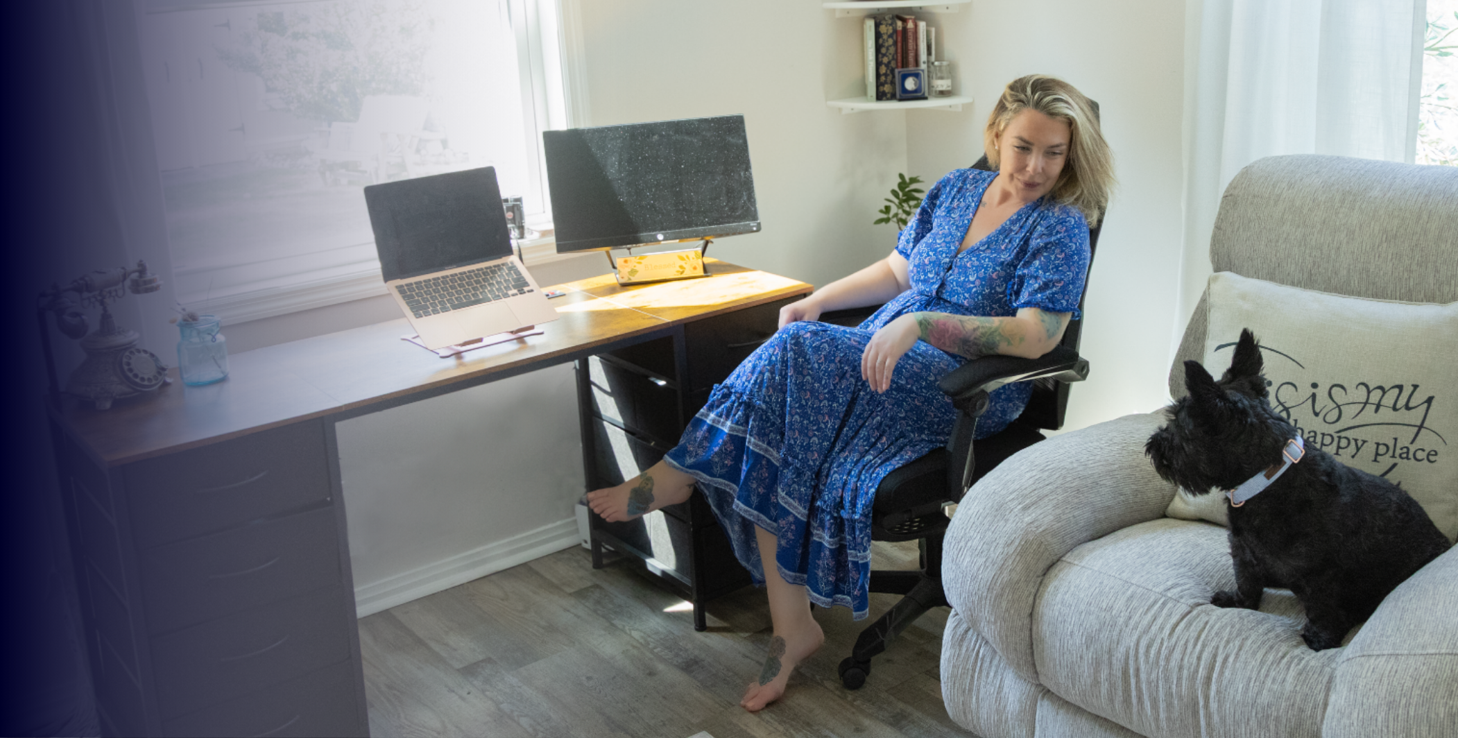 Woman in a blue dress sitting in an office chair in a home office, with a small black dog sitting on the couch nearby. The room is well-lit, with computers, books, and decorative items on the desk and shelves.