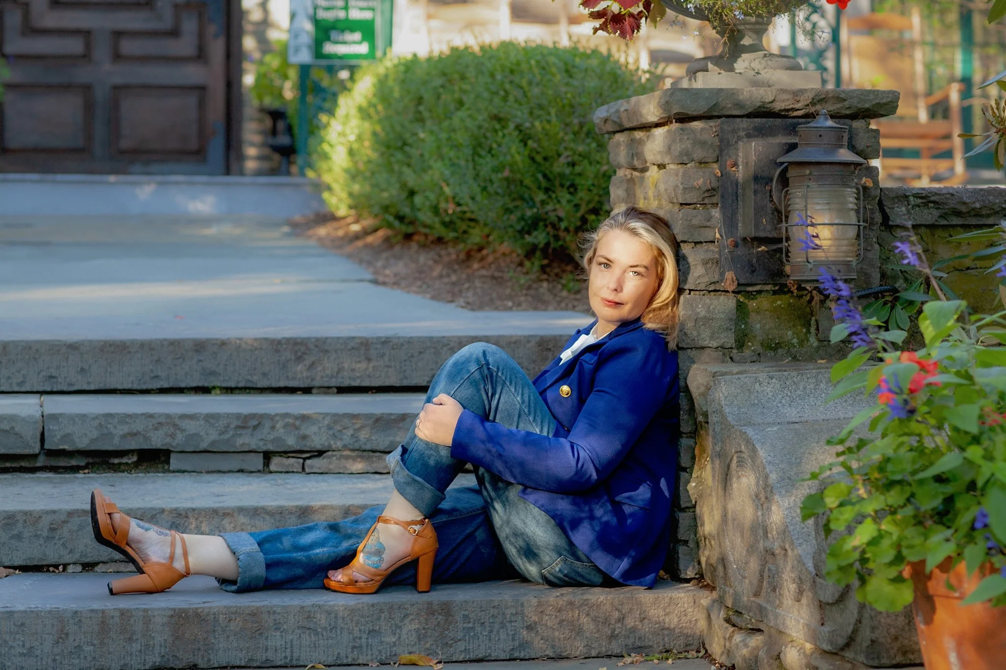 A woman with blonde hair wearing a blue blazer, jeans, and tan high heels, sitting on stone steps next to a stone wall with landscaped bushes and plants around her.