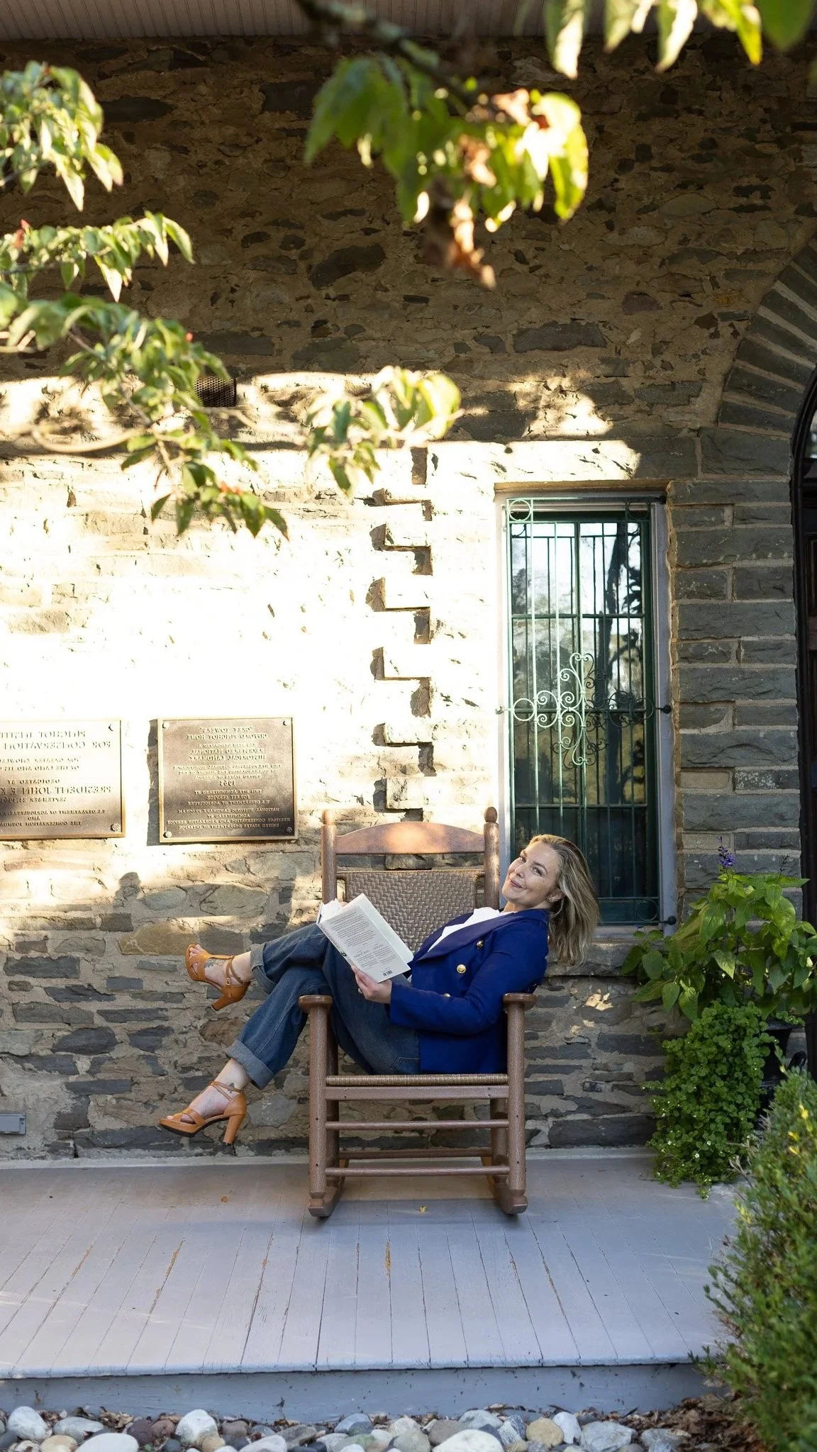 Woman sitting relaxed on a wooden chair outdoors, reading a book, next to a stone wall with plaques and a window, surrounded by greenery.