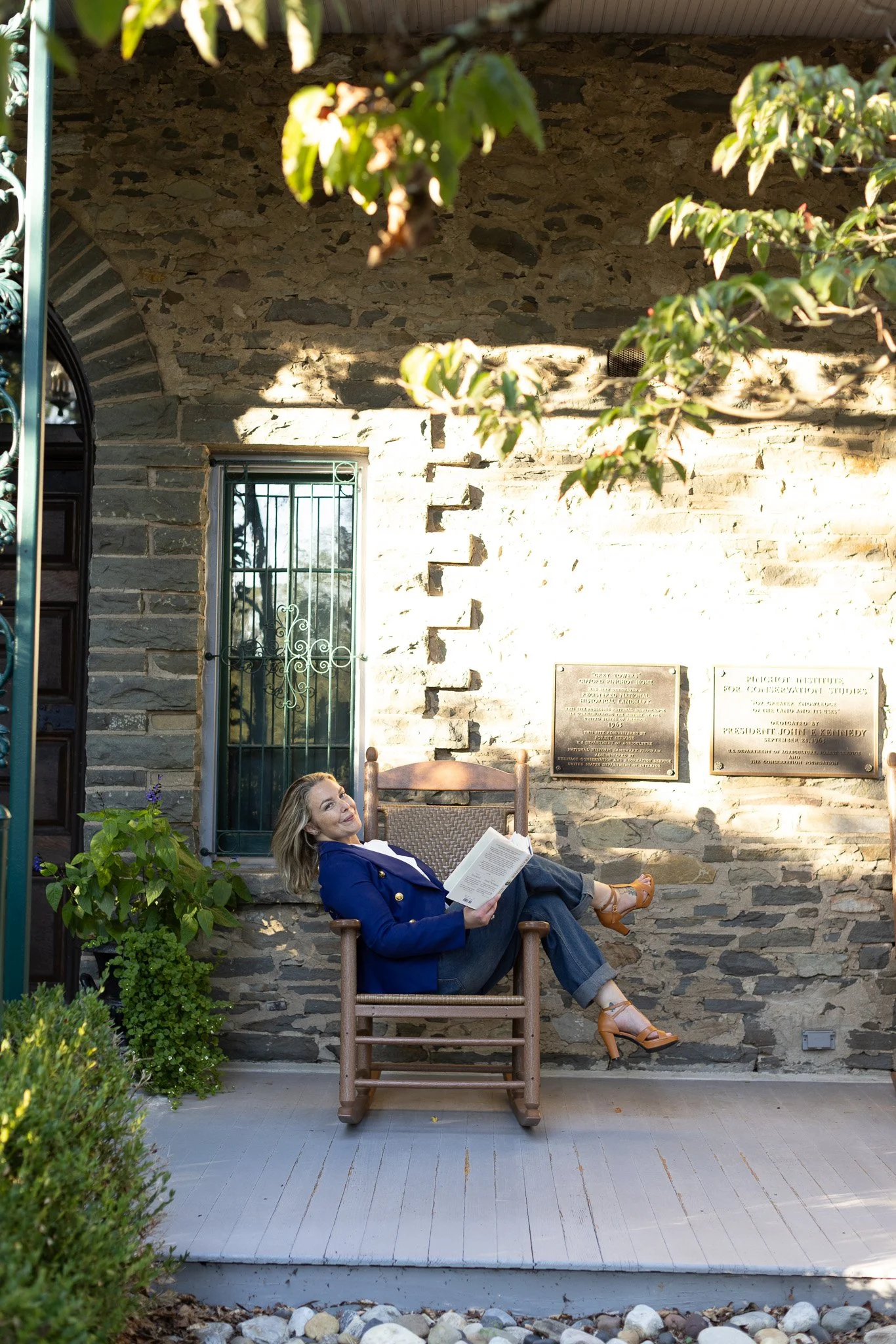 Woman sitting in a wooden chair on a porch, reading a book, with a stone wall and plaques in the background.