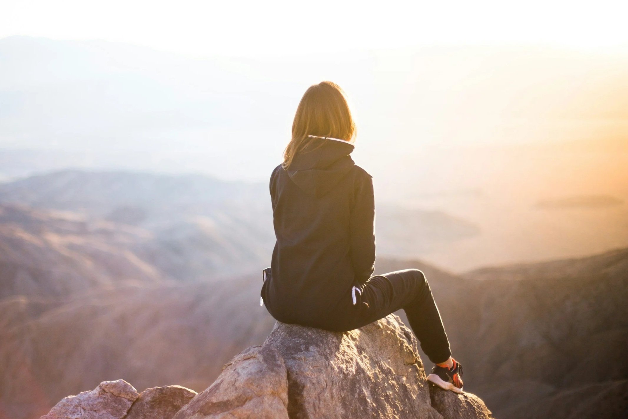 Person sitting on a rock ledge gazing at a mountainous landscape during sunset.